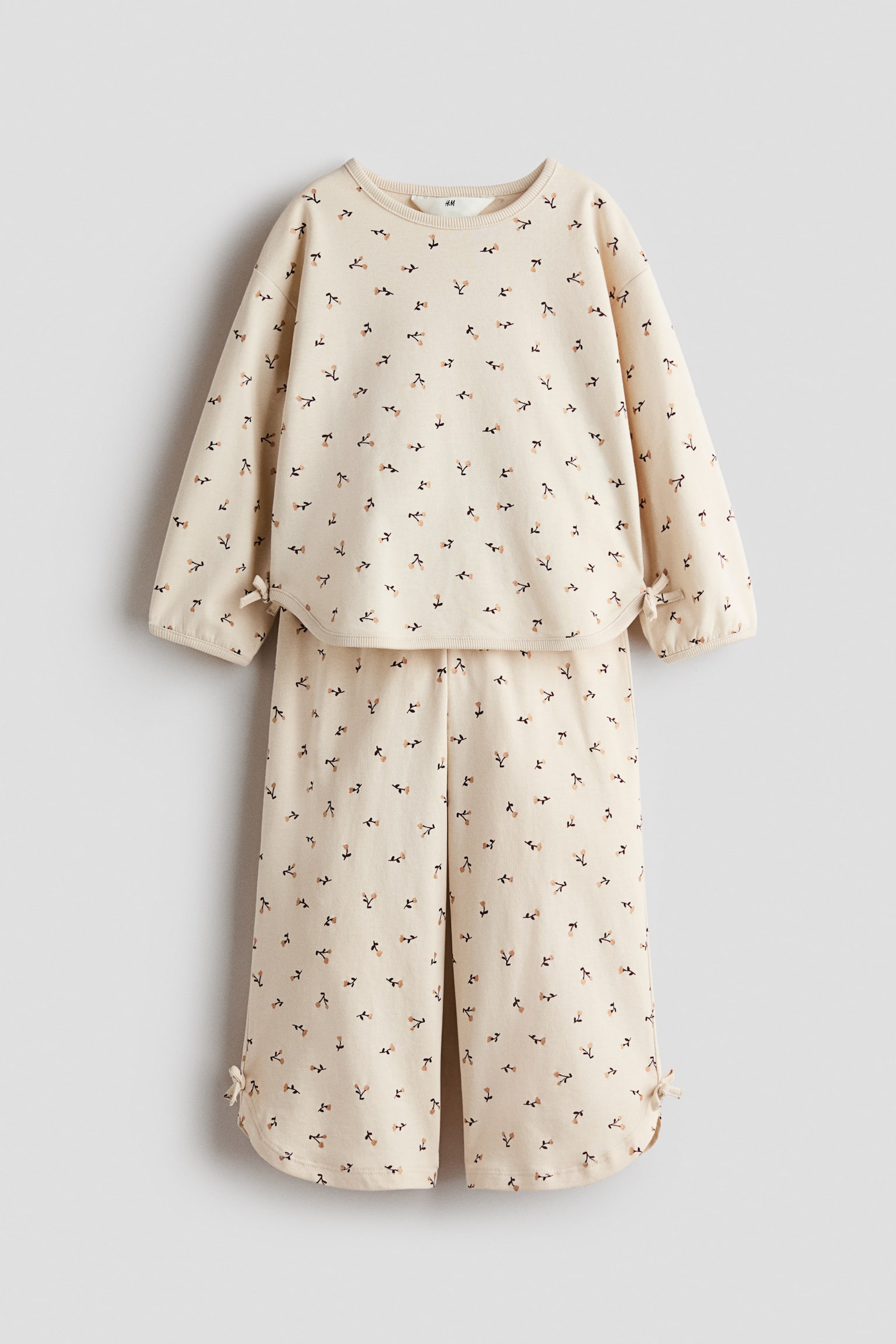 A front-facing view displays a matching cream-coloured two-piece set featuring a small floral pattern throughout. The long-sleeve top is designed with dropped shoulders, a ribbed crew neck, and a rounded hem with small decorative bows at the side vents, while the matching wide-leg trousers show similar bow-detailed side vents at the bottom hems.