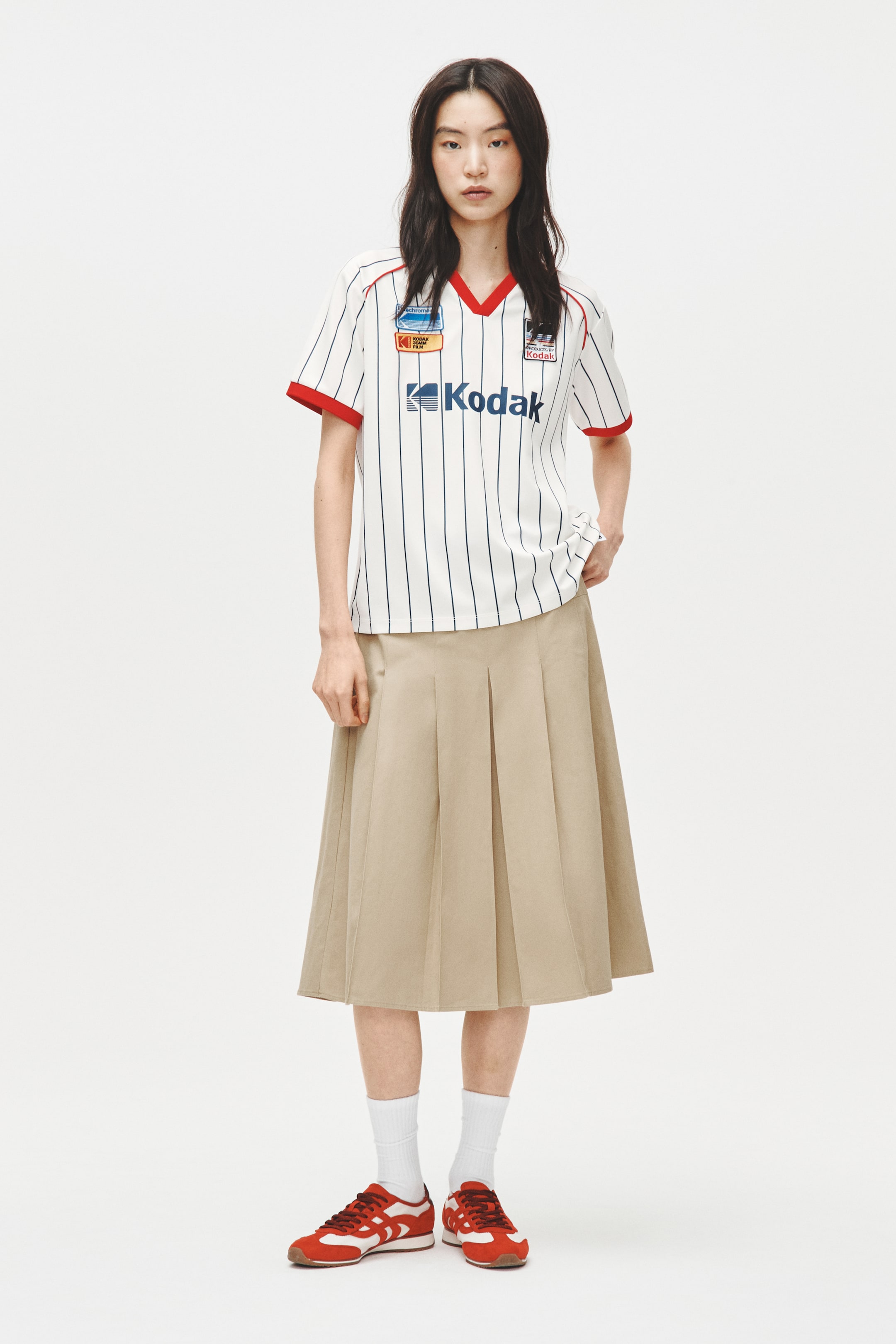 View larger image: A complete outfit is showcased, featuring a white short-sleeve shirt with thin navy vertical stripes, a red V-neck collar, and red sleeve cuffs, displaying a blue "Kodak" logo and various brand patches on the front. The ensemble is finished with a pleated khaki midi skirt and red sneakers with white accents, paired with white ankle socks.