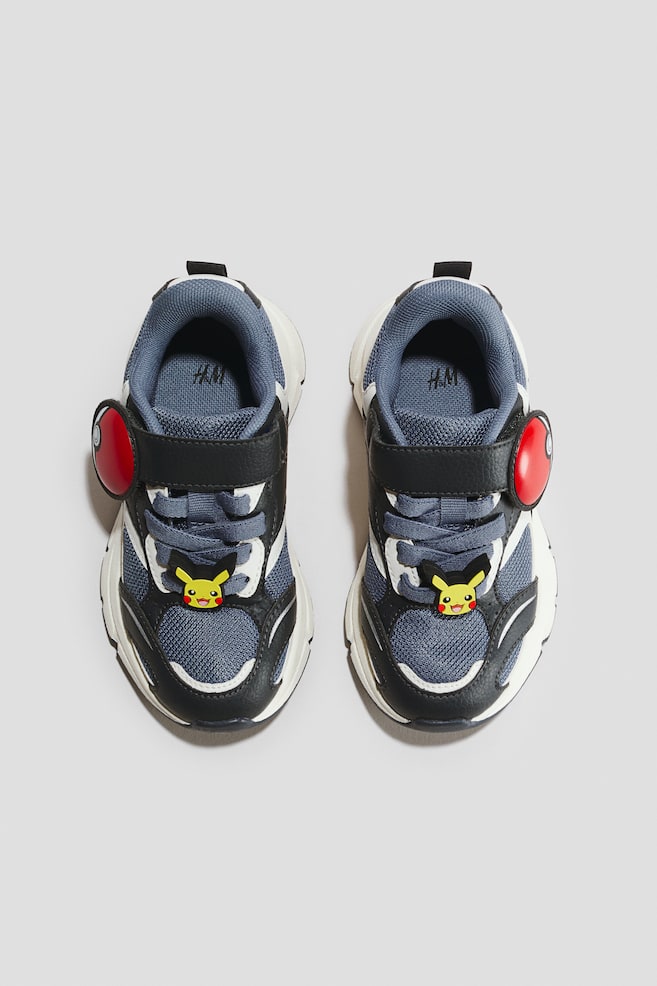Ein Paar Kindersneaker zeigt graue Mesh-Obermaterialien mit schwarzen Kunstleder-Overlays und klobigen weißen Sohlen, die nach vorne gerichtet sind. Jeder Schuh ist mit einem schwarzen Klettverschluss mit einem roten und weißen Pokéball-Emblem ausgestattet, und ein gelber Pikachu-Charme ziert die grauen elastischen Schnürsenkel. - 1