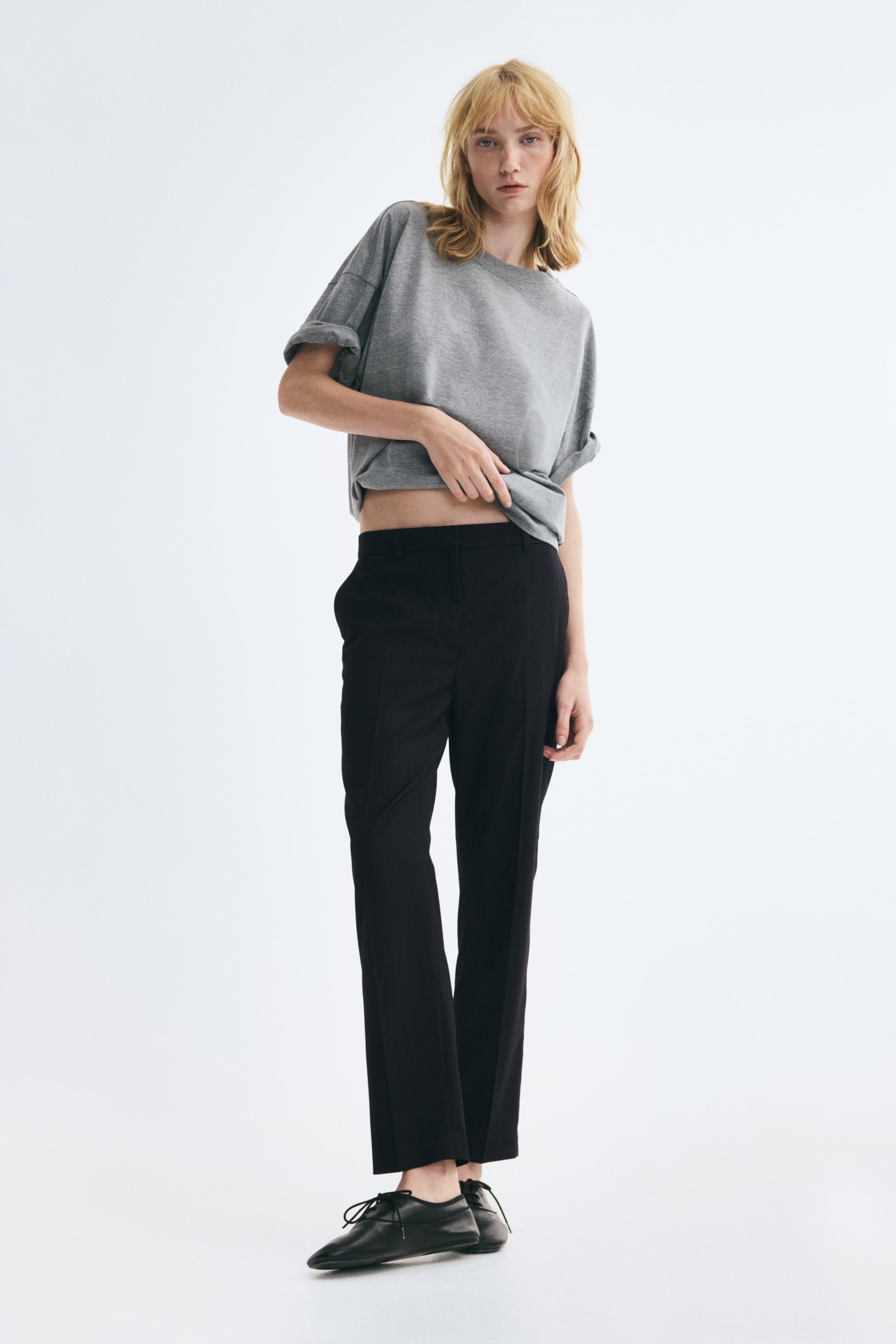 Größeres Bild anzeigen: Schwarze, knöchellange Hose aus Webstoff, getragen vom Model, mit schmal zulaufenden Beinen und sichtbaren Bügelfalten vorne. Das Model hebt den Saum eines grauen T-Shirts an, um den flachen Bund der Hose zu zeigen, die mit schwarzen flachen Schuhen kombiniert ist.
