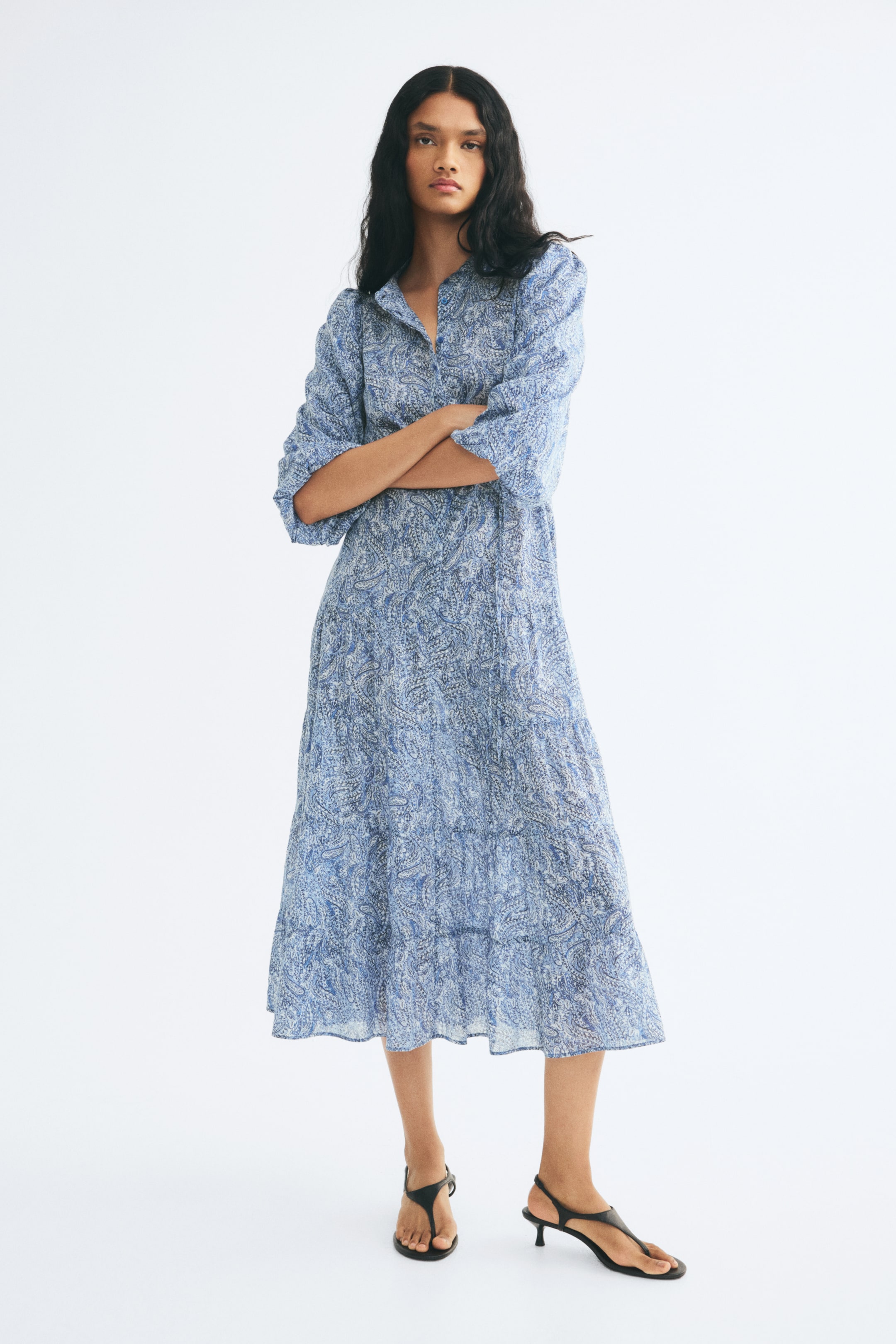 View larger image: A long, relaxed-fit dress with a blue paisley pattern is being worn by the model, who is standing with her arms crossed. The dress features a band collar, three-quarter-length puff sleeves, and a tiered midi skirt, styled with black kitten-heel sandals.