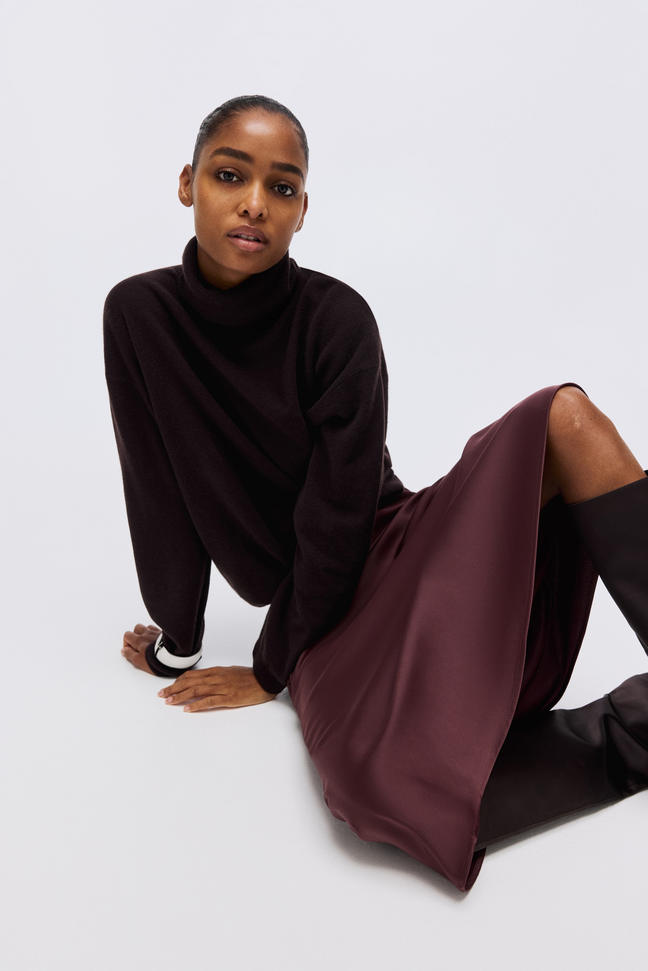 Voir une image plus grande: Un chandail à col roulé brun foncé présente une coupe décontractée et des manches longues. Il est associé à une jupe en satin bordeaux qui flotte autour des jambes, mettant en valeur sa texture lustrée et son tombé fluide.