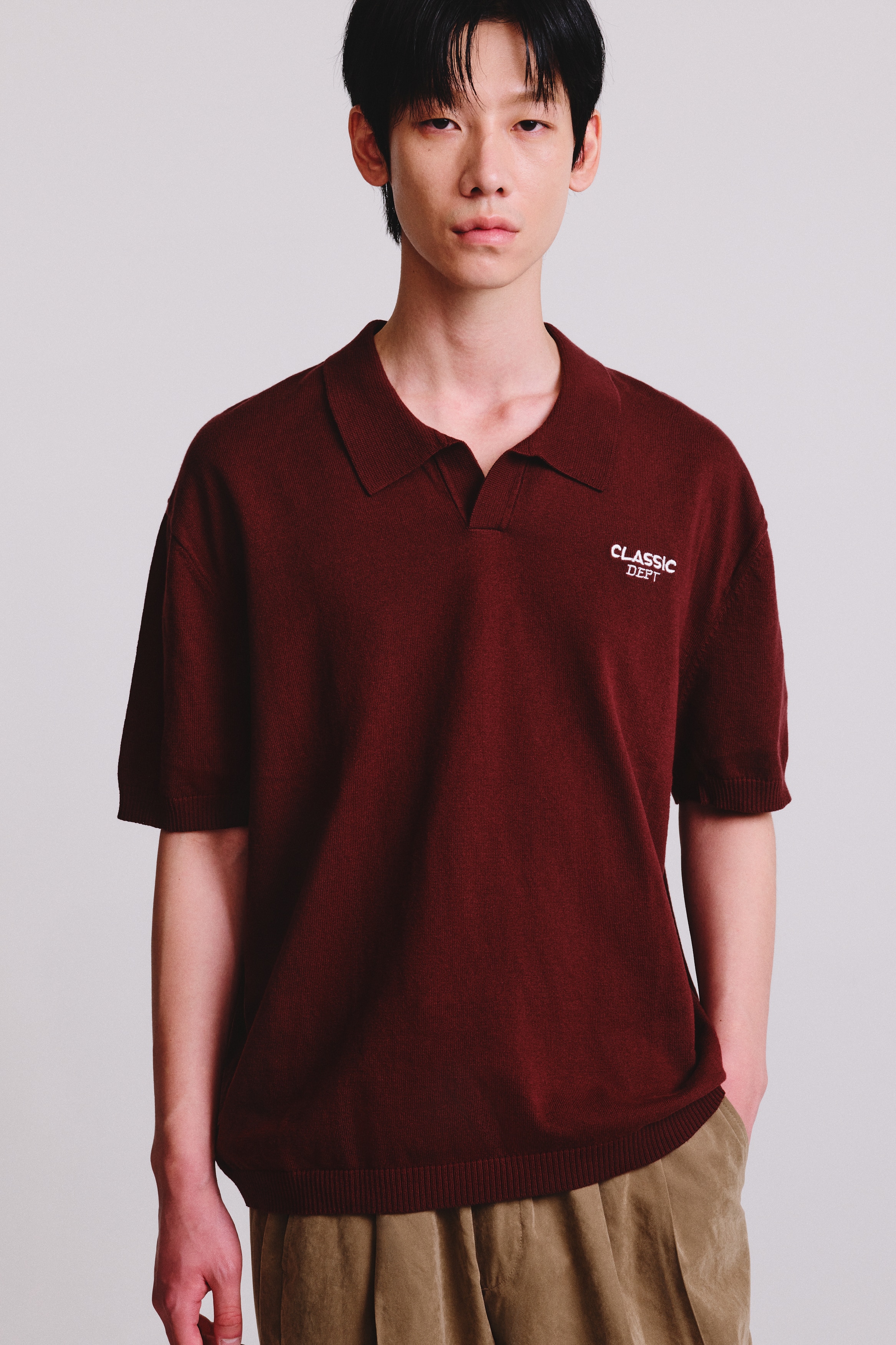 A burgundy knit polo shirt is shown from the front, featuring a flat-knit collar with a buttonless V-neckline. The shirt has short sleeves with ribbed cuffs, a ribbed hem, and white embroidered text reading "CLASSIC DEPT" on the left chest.