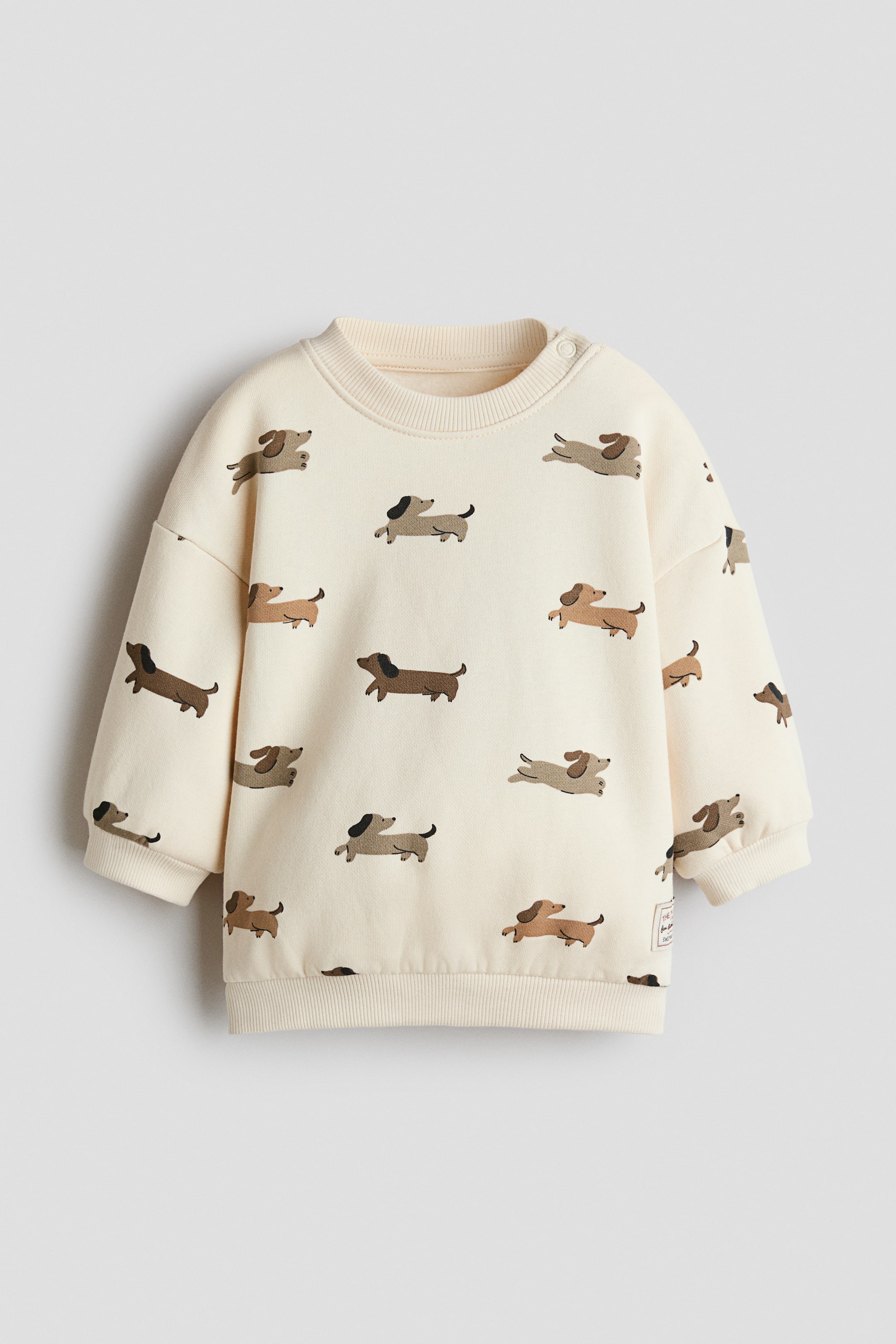 View larger image: A cream-coloured sweatshirt features a repeated pattern of brown and black dachshunds across the front. It is designed with a ribbed crew neck, two snap buttons on the left shoulder, ribbed cuffs, and a ribbed hem.