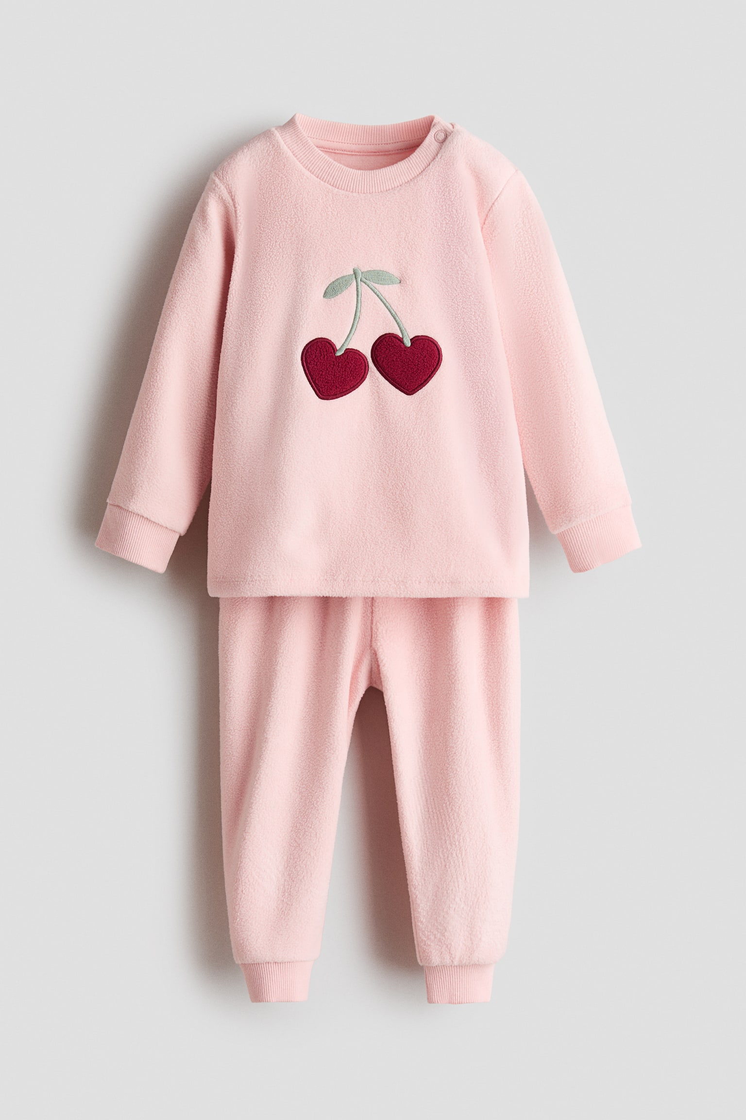 Un ensemble de pyjama en polaire rose clair est prÃ©sentÃ© de face, comprenant un haut Ã  manches longues et un pantalon assorti. Le haut arbore une applique centrale reprÃ©sentant deux cerises bordeaux en forme de cÅur avec de petites tiges vert clair, ainsi quâun col rond cÃ´telÃ© et des poignets cÃ´telÃ©s. Le pantalon assorti complÃ¨te lâensemble avec des bas de jambes cÃ´telÃ©s aux chevilles.