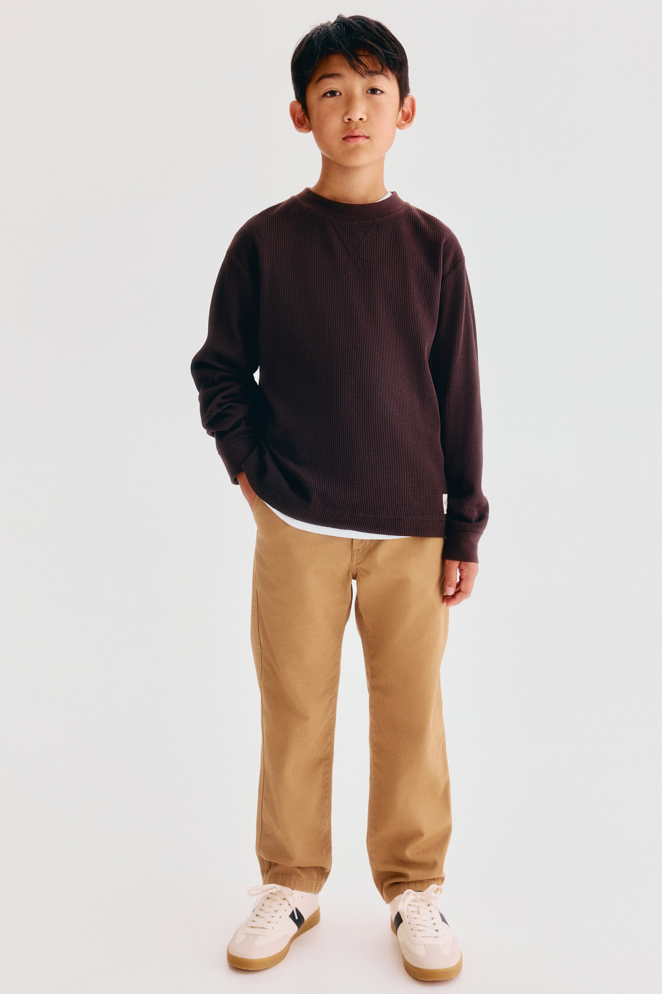 Grotere afbeelding bekijken: Chino’s in katoenen twill zijn van voren getoond op een model, met een rechte pijpsnit in een warme beige tint. De broek is gestyled met een donkerbruine, getextureerde sweatshirt met lange mouwen en witte sneakers, waarbij één hand in een diagonale steekzak aan de voorkant rust.