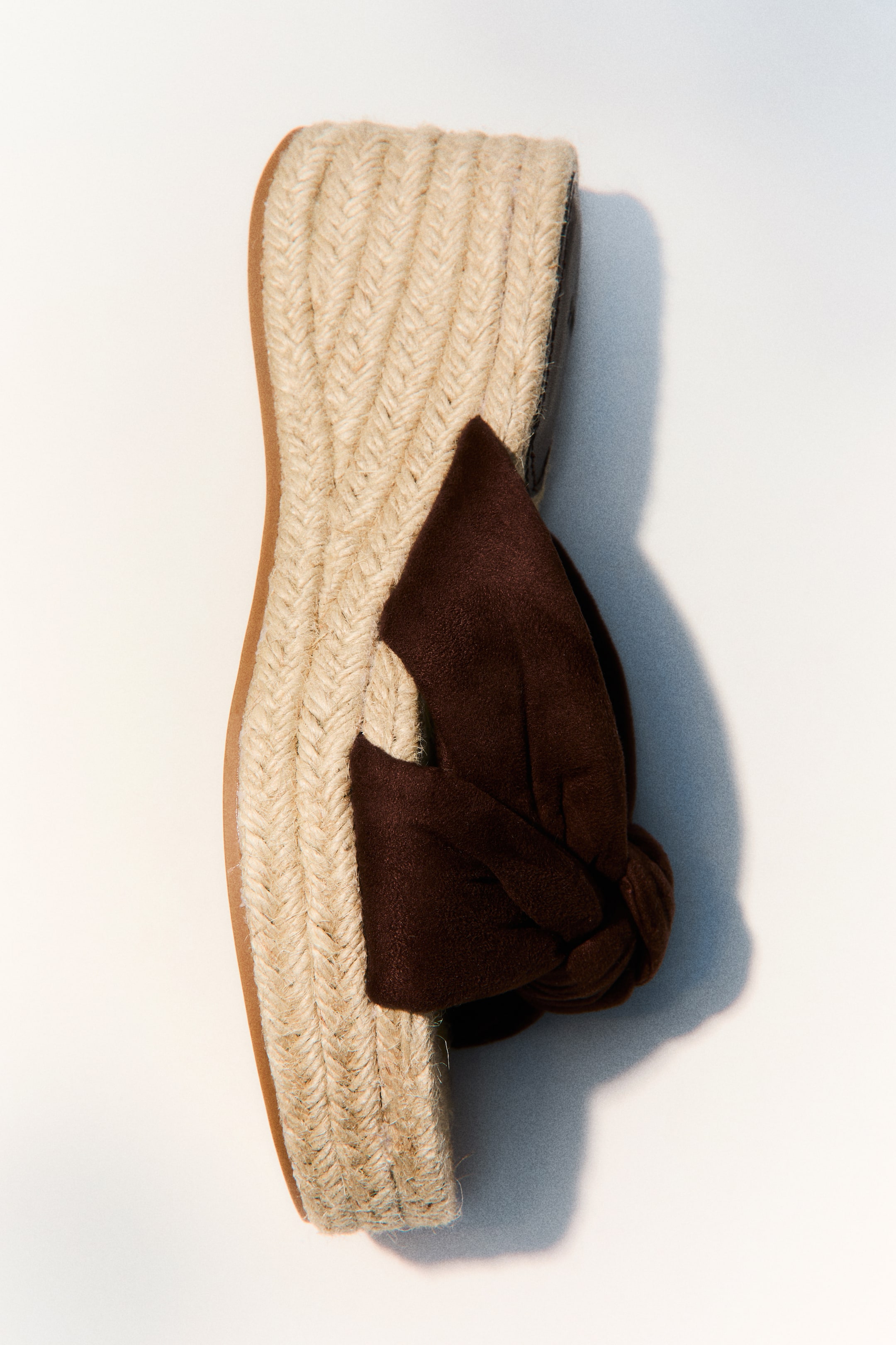 View larger image: A single espadrille wedge sandal is positioned diagonally, highlighting its light beige woven jute sole with a herringbone pattern. A dark brown fabric strap is elegantly knotted across the top of the sole.