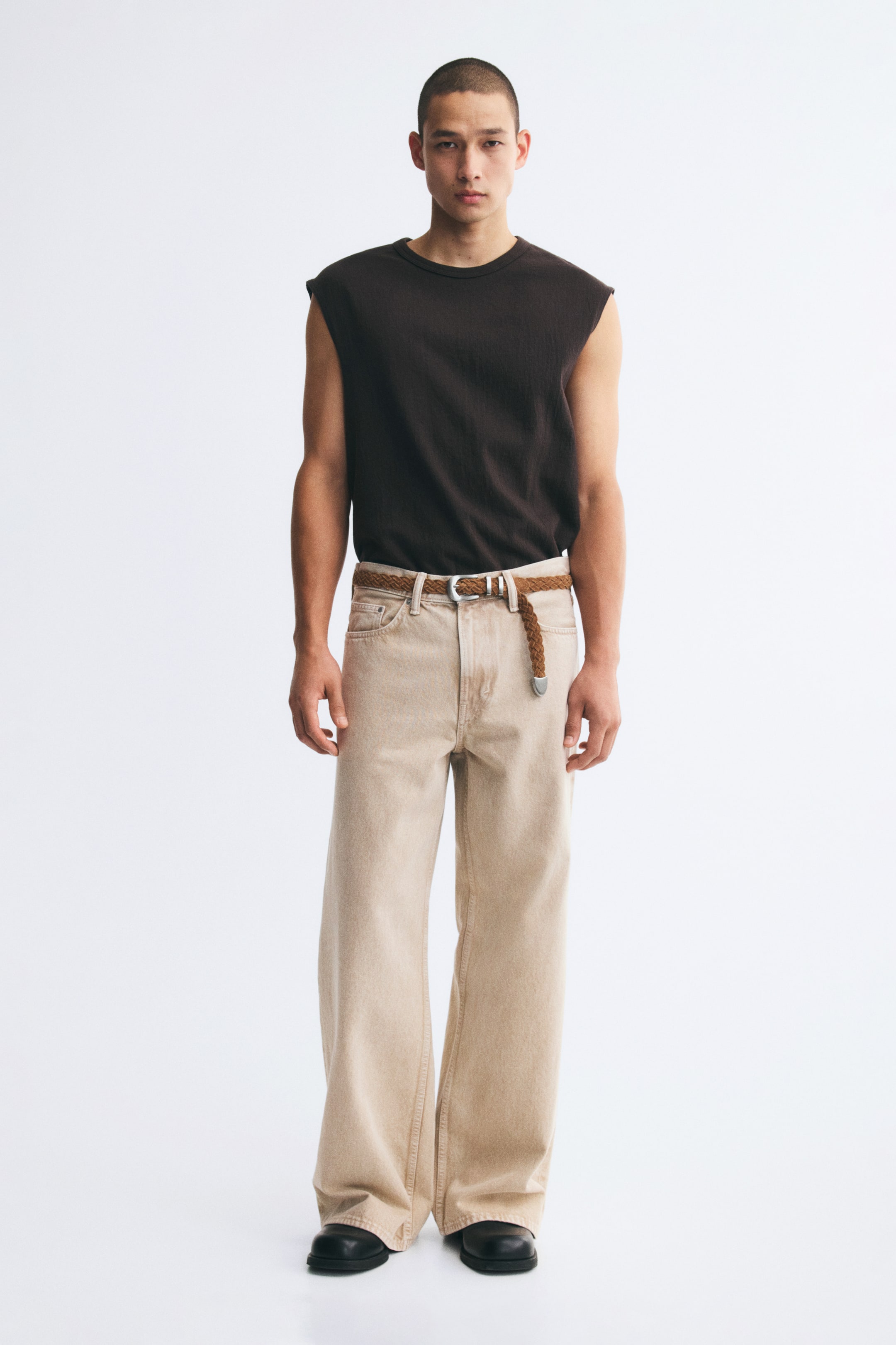 View larger image: Light beige wide-leg denim jeans with a loose fit and a regular waist are worn by the model. The jeans are styled with a braided brown belt and a dark brown sleeveless top tucked into the waistband.