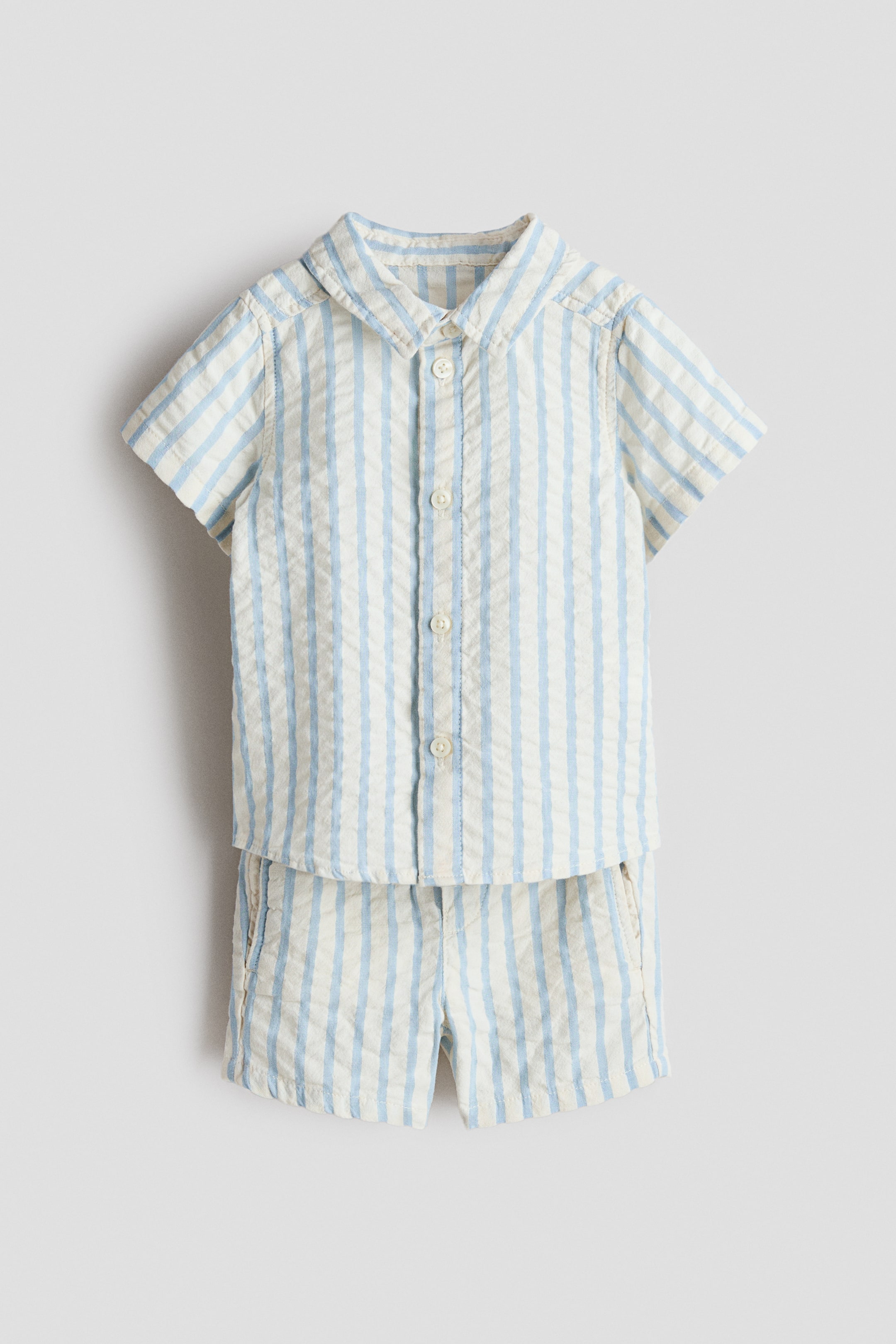 View larger image: A baby's two-piece outfit is presenting FRONT, featuring a short-sleeved shirt with a collar, a full button-down front, and light buttons. Both the shirt and matching shorts are displaying light blue and off-white vertical stripes on a textured fabric, with the shorts also showing side pockets.