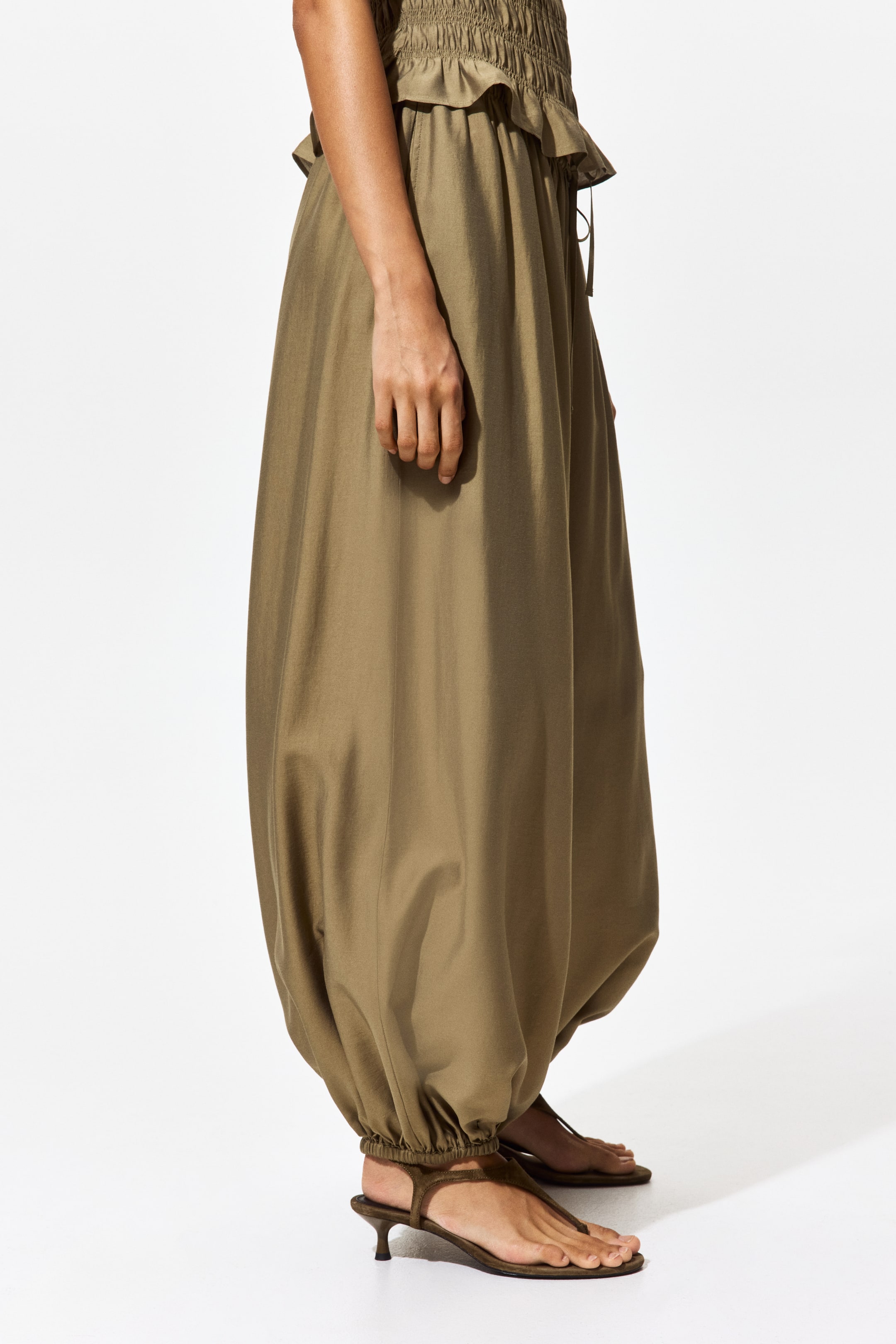 View larger image: An olive green garment, showcasing a smocked waist with a ruffled trim and voluminous legs, is being worn from a side angle. The legs gather at the ankles, resting just above a pair of sandals.