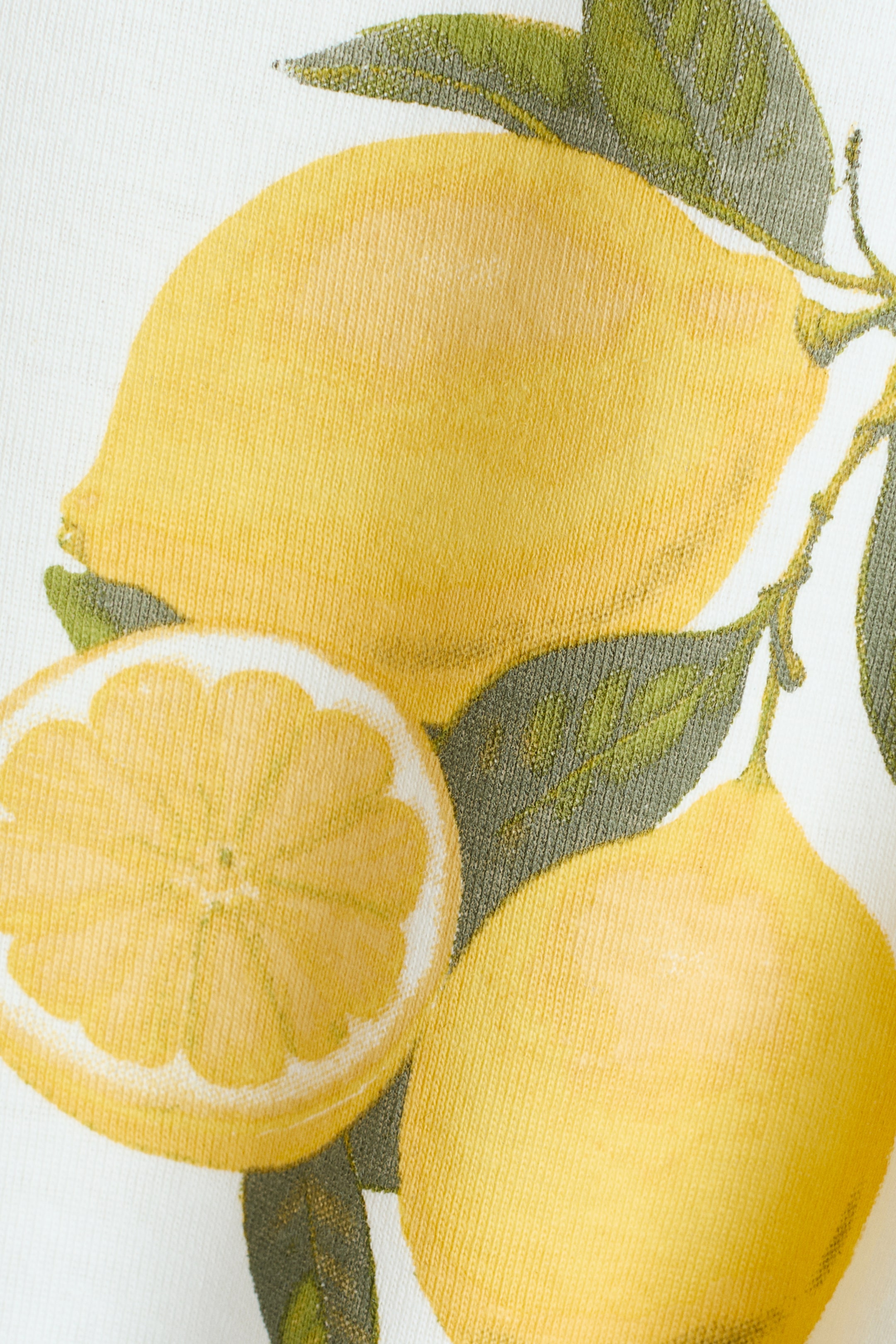 Grotere afbeelding bekijken: Een T-shirt met een close-up van zijn zachte, bedrukte jersey stof. Het toont een levendig patroon van hele gele citroenen, een gesneden citroen en groene bladeren.