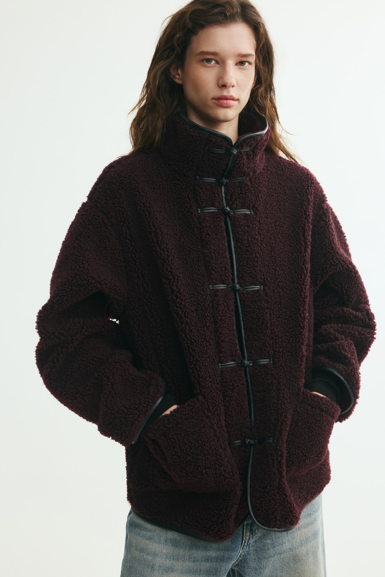 An oversized burgundy teddy jacket with a high funnel collar, black coated-fabric trim, and front knot closures is worn by the model. The model is standing with hands resting in the front patch pockets, styled with light blue jeans. - 1