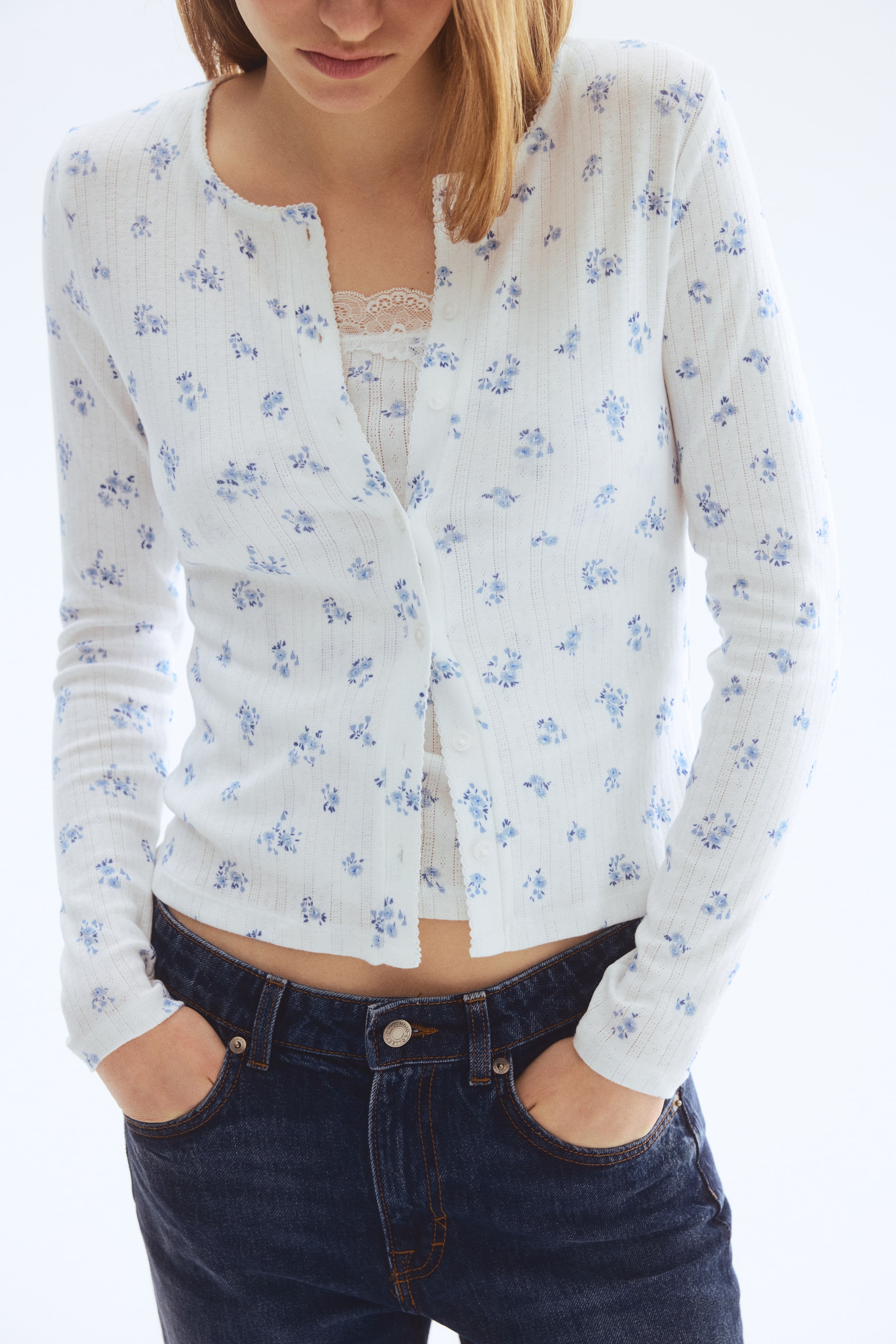 View larger image: A white ribbed cardigan adorned with a blue floral print features a round neckline and a button-front closure. It's styled cropped over a white lace-trimmed camisole and paired with dark jeans.