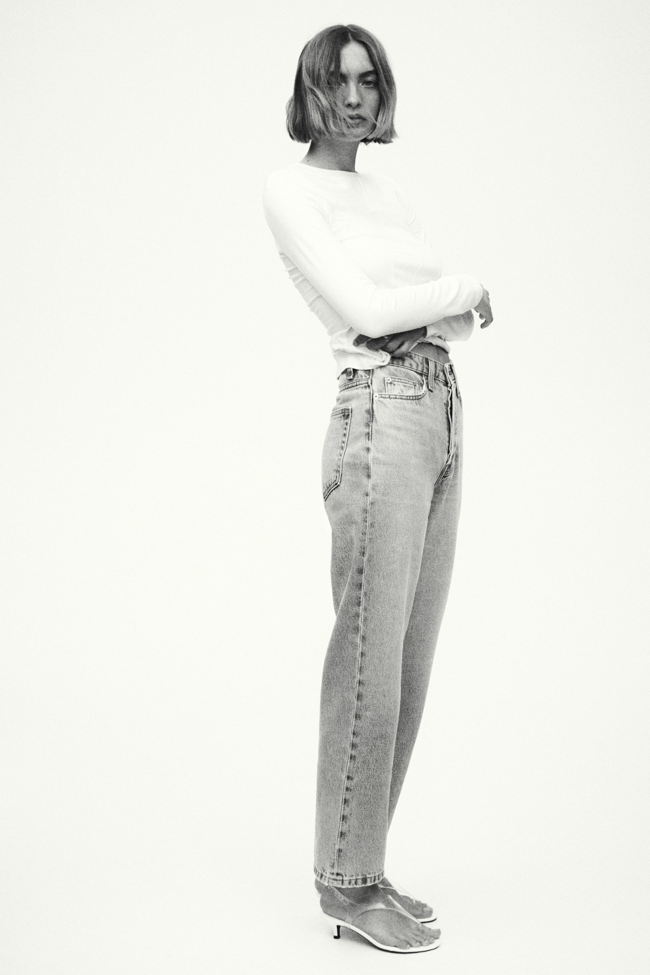 Größeres Bild ansehen: Hell gewaschene, hoch taillierte 5-Pocket-Jeans mit Knopfverschluss werden vom Model getragen und zeichnen sich durch ein gerades Bein aus, das bis zu einer knöchellangen Höhe über klaren Absatzschuhen fällt. Das Model steht mit einer Hand in der Nähe des Bunds, und die Jeans sind mit einem eingesteckten langärmeligen Oberteil gestylt.