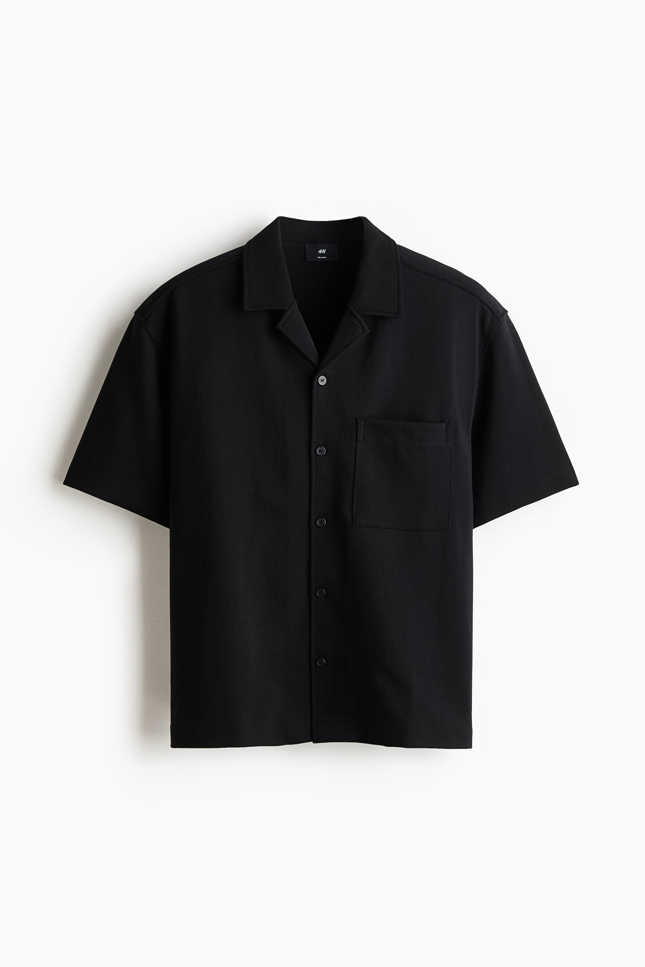 Ver imagen más grande: Una camisa negra de manga corta se presenta de frente, con un cuello de solapa, una botonadura completa con botones oscuros y un único bolsillo de parche en el pecho izquierdo. La tela muestra un sutil tejido texturizado.