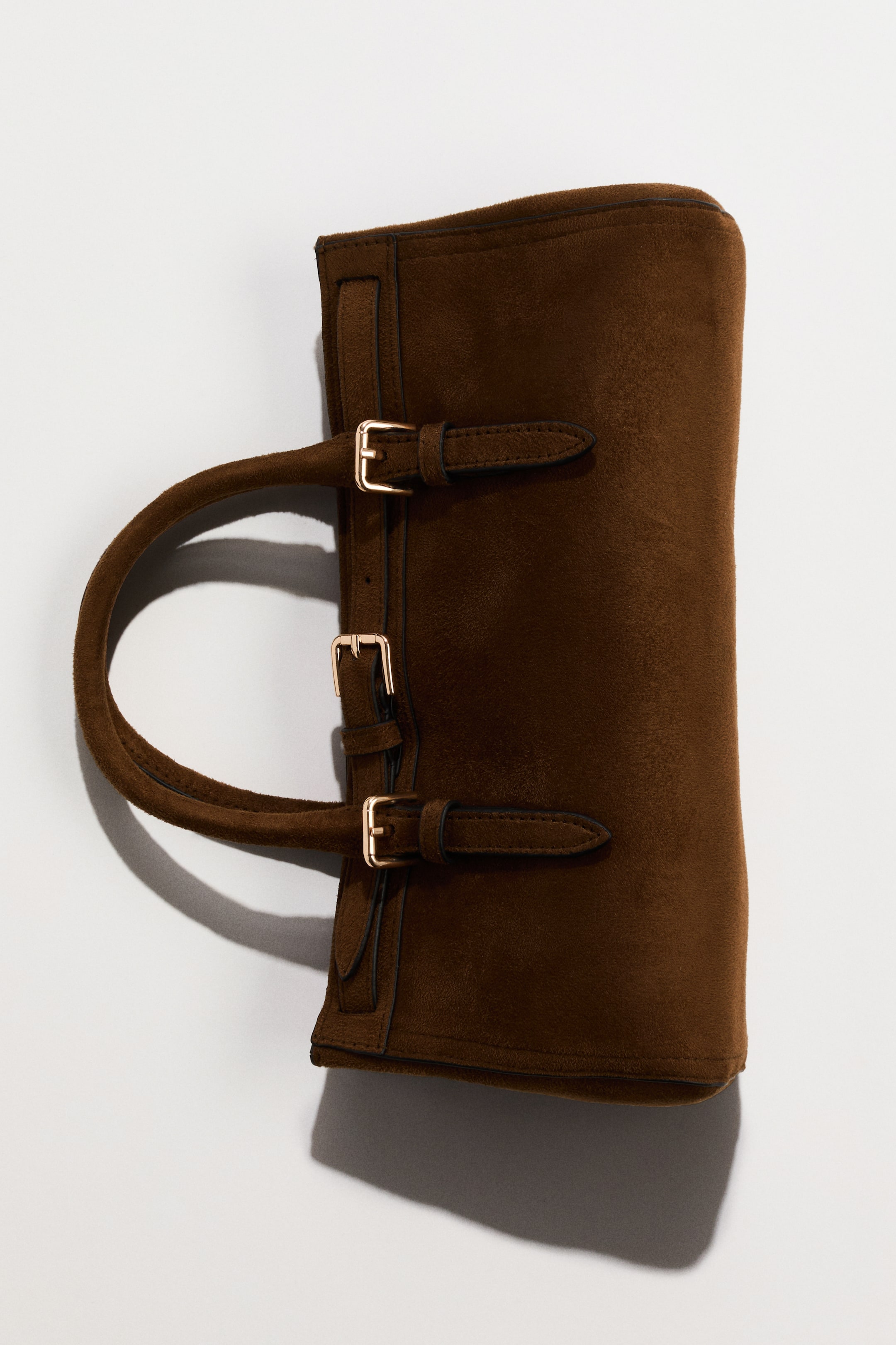 View larger image: A brown suede bag is positioned vertically, showcasing its front and side. It features two elegantly curved handles extending from the top edge and three gold-toned buckles with straps running vertically down its front.