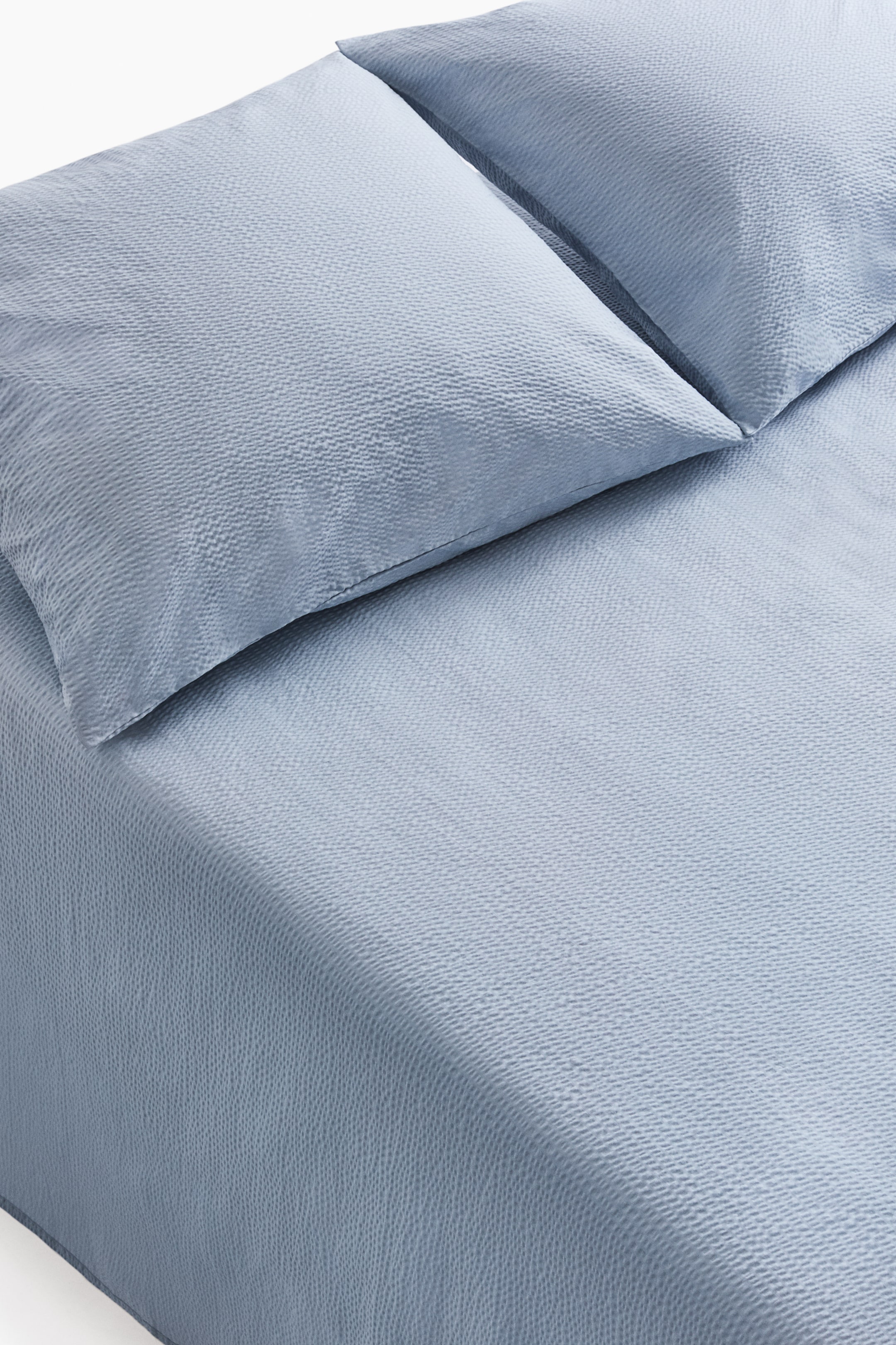 View larger image: Light blue bed linen is showcased, comprising two pillows and a duvet cover. Each piece exhibits a gentle, textured, rippled surface, with one pillow slightly overlapping the other.