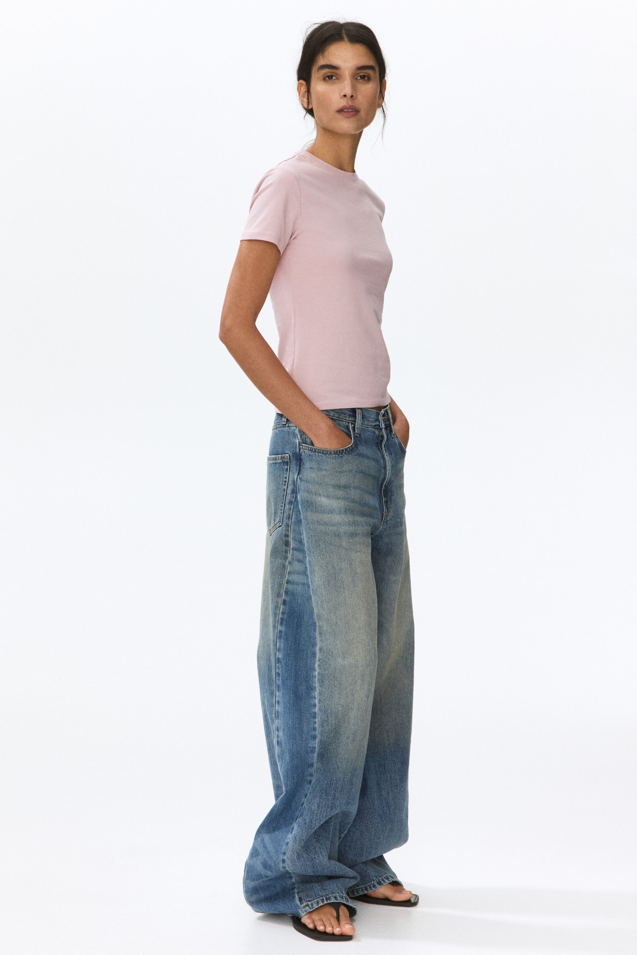 View larger image: A pink crew neck t-shirt is depicted from a side angle, showcasing short sleeves and a fitted silhouette. It is styled tucked into jeans, highlighting its length and fit.