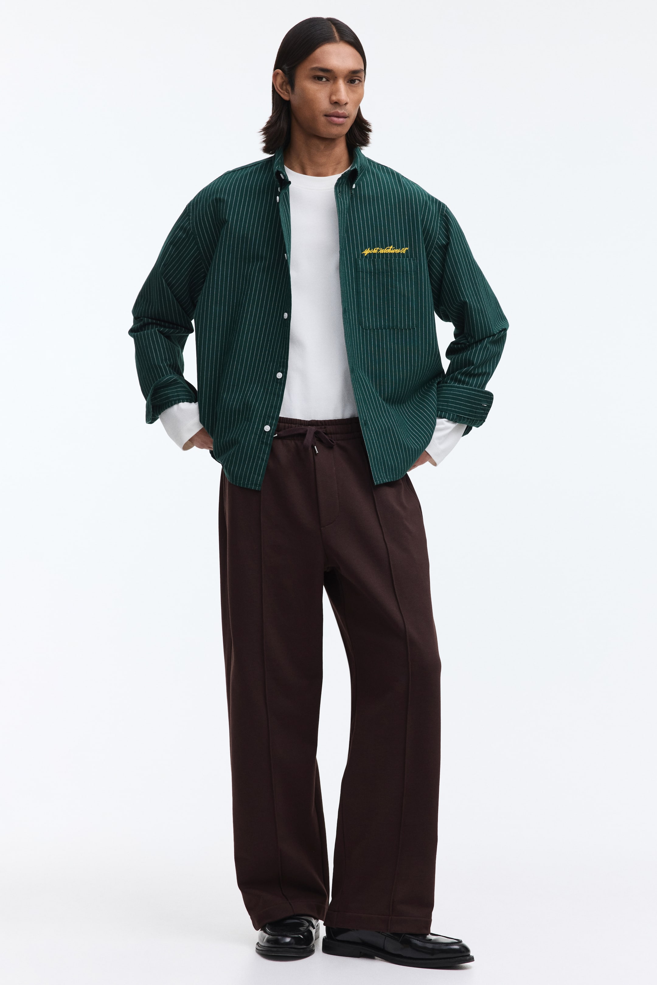 이미지 확대 보기: 이 의상은 얇은 흰색 세로 줄무늬와 노란색 가슴 자수가 있는 다크 그린 긴소매 셔츠를 흰색 긴소매 티셔츠 위에 오픈 스타일로 연출한 것입니다. 또한, 허리에 드로스트링이 있는 다크 브라운 와이드 레그 바지와 각 다리의 세로 솔기가 특징이며, 블랙 광택 로퍼로 마무리되었습니다.