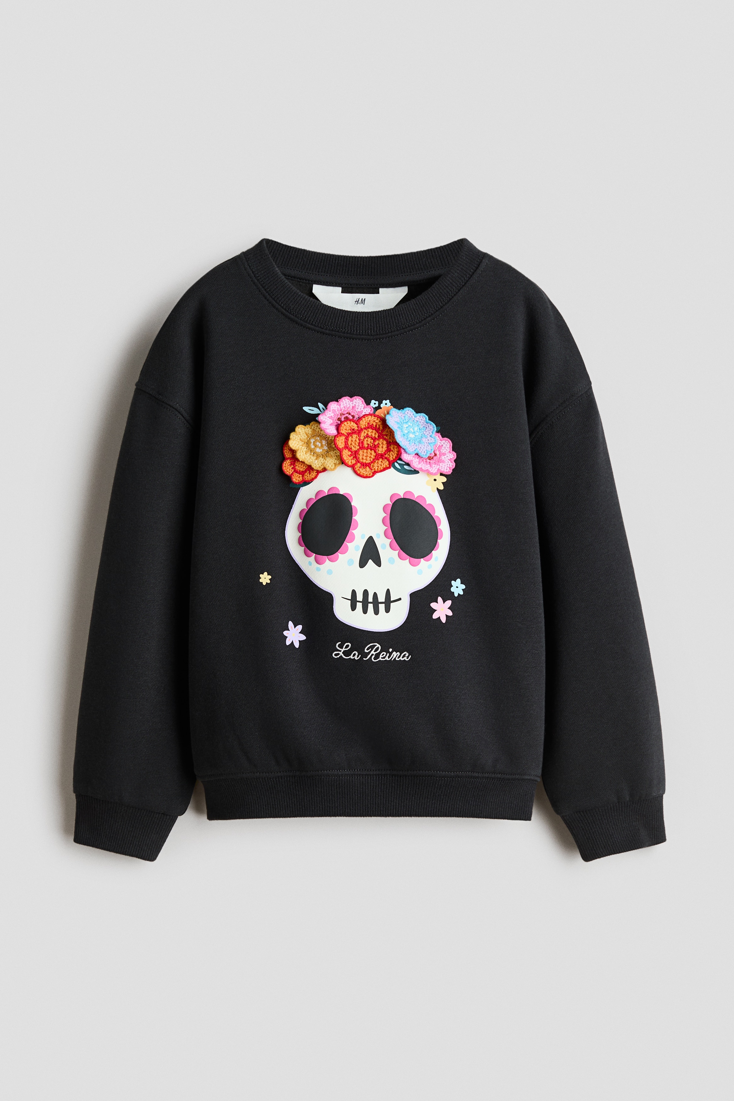 A black crewneck fleece sweater is displaying a front-facing sugar skull graphic adorned with textured, colorful crocheted flower appliques. The design is featuring small printed pastel flowers around the skull and the white cursive text "La Reina" at the bottom, with white long-sleeve cuffs peeking out from underneath the ribbed cuffs.