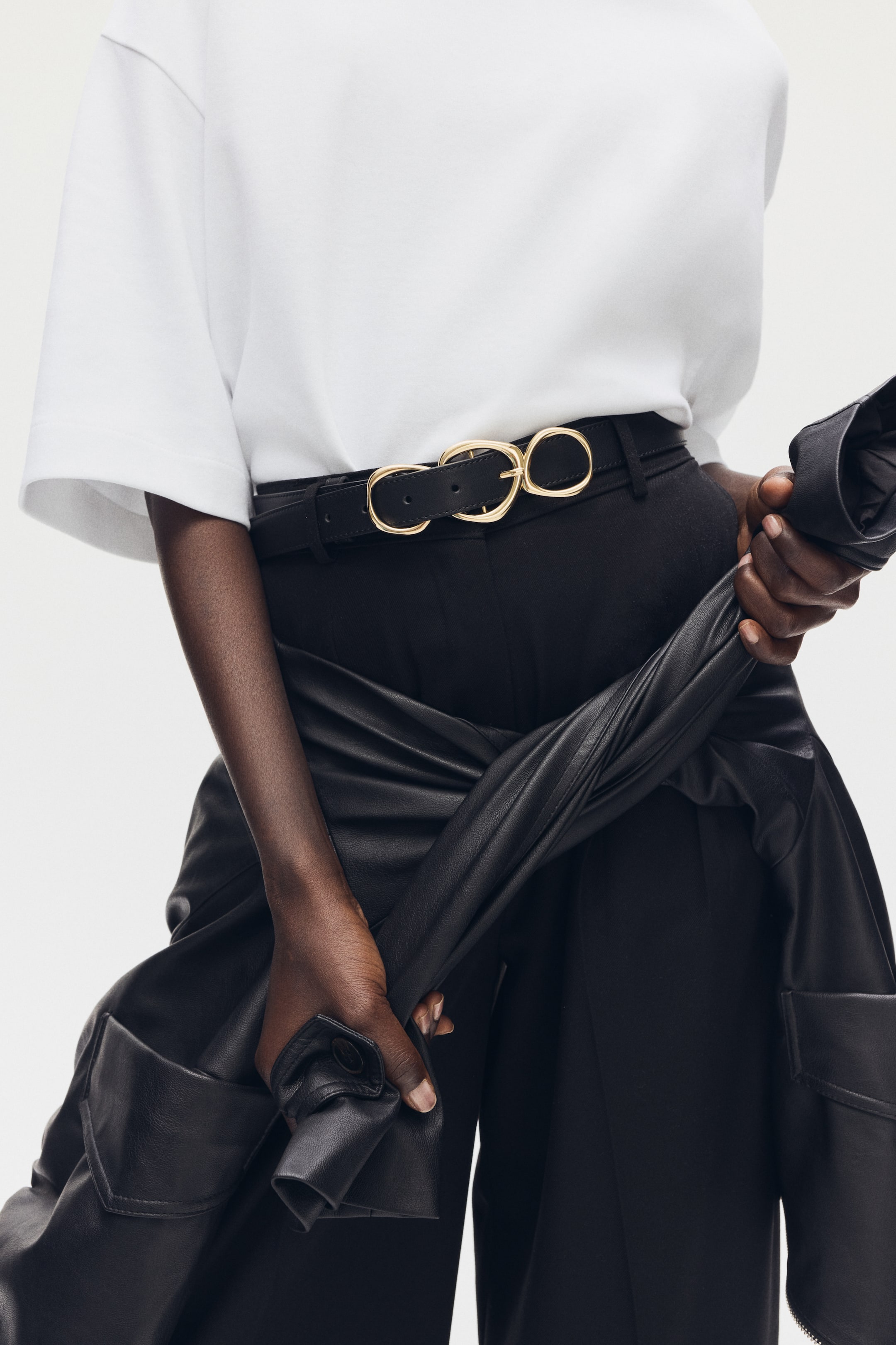 View larger image: A black leather-look belt featuring a gold-toned, abstract interlocking buckle is styled around the waist of black trousers, paired with a white top.