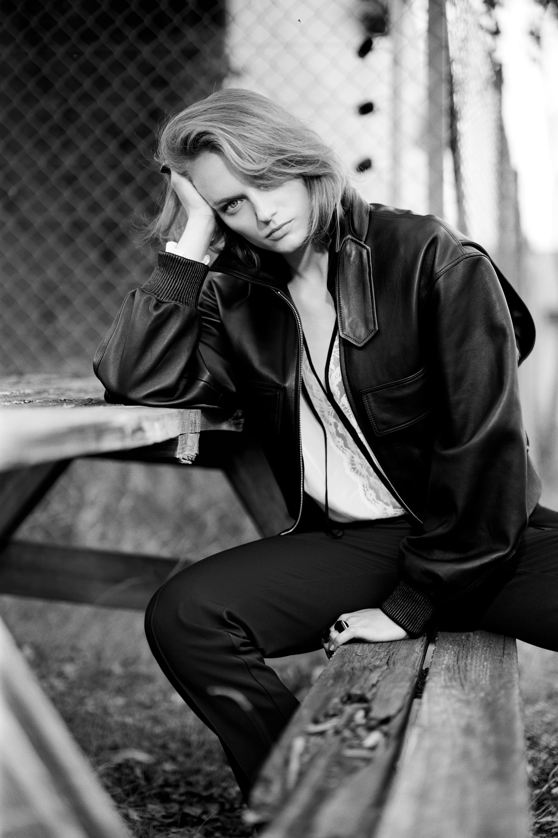 Agrandir l'image: Une veste bomber en cuir noir, dotée de poignets côtelés, de poches avant et d’un col classique, est portée par le mannequin. La veste est stylisée ouverte sur un haut clair bordé de dentelle et un pantalon foncé, tandis que le mannequin est assis sur un banc en bois.