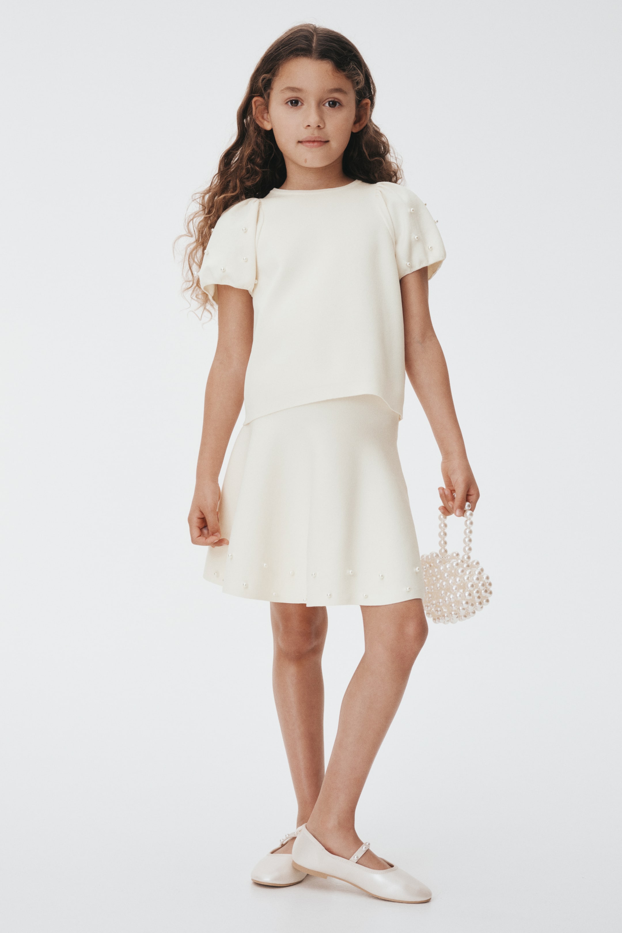 הצגת תמונה גדולה יותר: בגד שני חלקים בצבע קרם מוצג, הכולל חולצה קצרה עם שרוולים תפוחים וצווארון עגול, וחצאית בגזרה A. פנינים פזורות מעטרות את שרוולי החולצה ושולי החצאית. תיק קטן עם חרוזי פנינה מוחזק ביד, ונעליים שטוחות בצבע קרם בסגנון מרי ג'יין עם רצועות מעוטרות פנינים ננעלות.