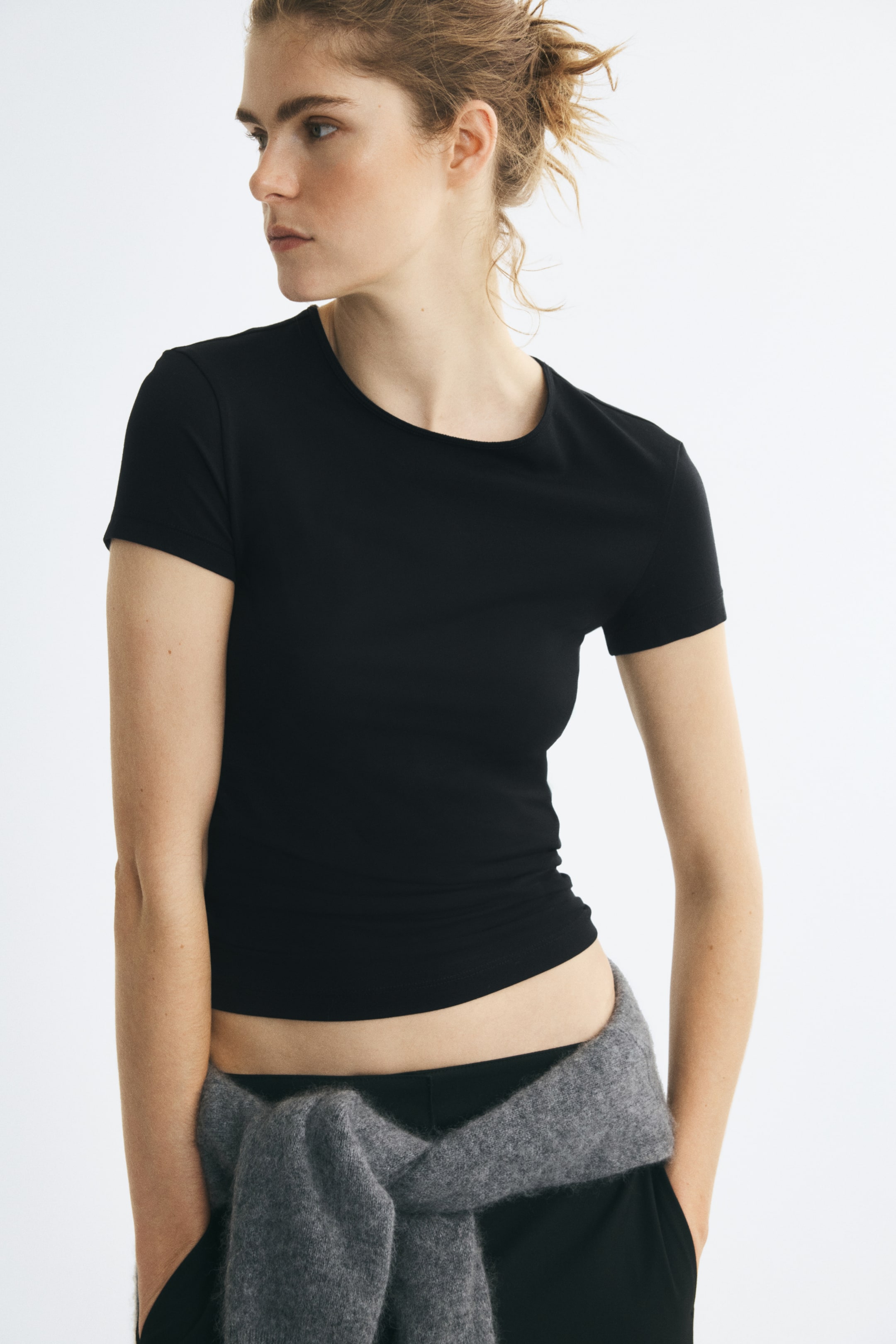 View larger image: The model is wearing a black short-sleeved crew-neck T-shirt. The fitted T-shirt is styled with black trousers and a grey knit sweater tied around the waist.