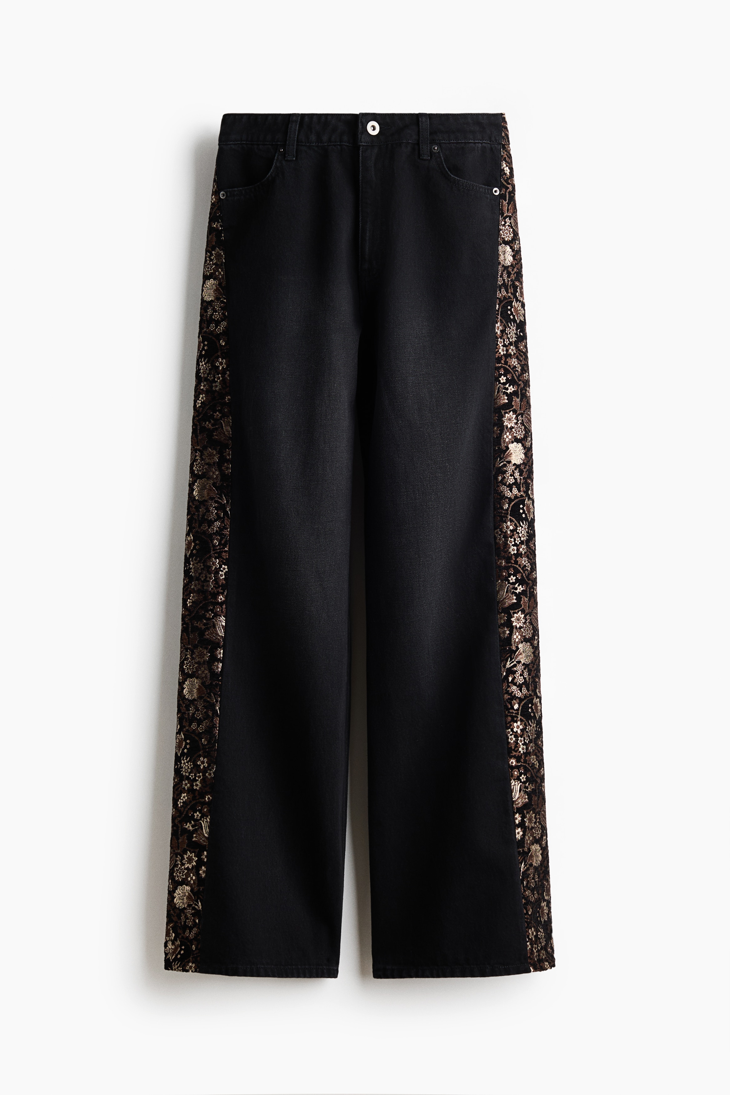 Loose-fit black denim jeans featuring a contrasting gold floral jacquard panel down the side are worn by the model. The low-waisted jeans are styled with a black leather belt with gold hardware and a long-sleeved black sweater.