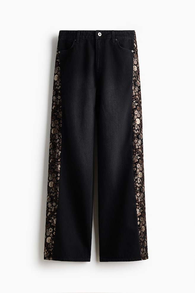 A pair of wide-leg black denim jeans is facing front, showcasing contrasting metallic-gold floral brocade panels running down both sides. The design is featuring a classic button-and-zip fly closure, belt loops, and curved front pockets. - 1