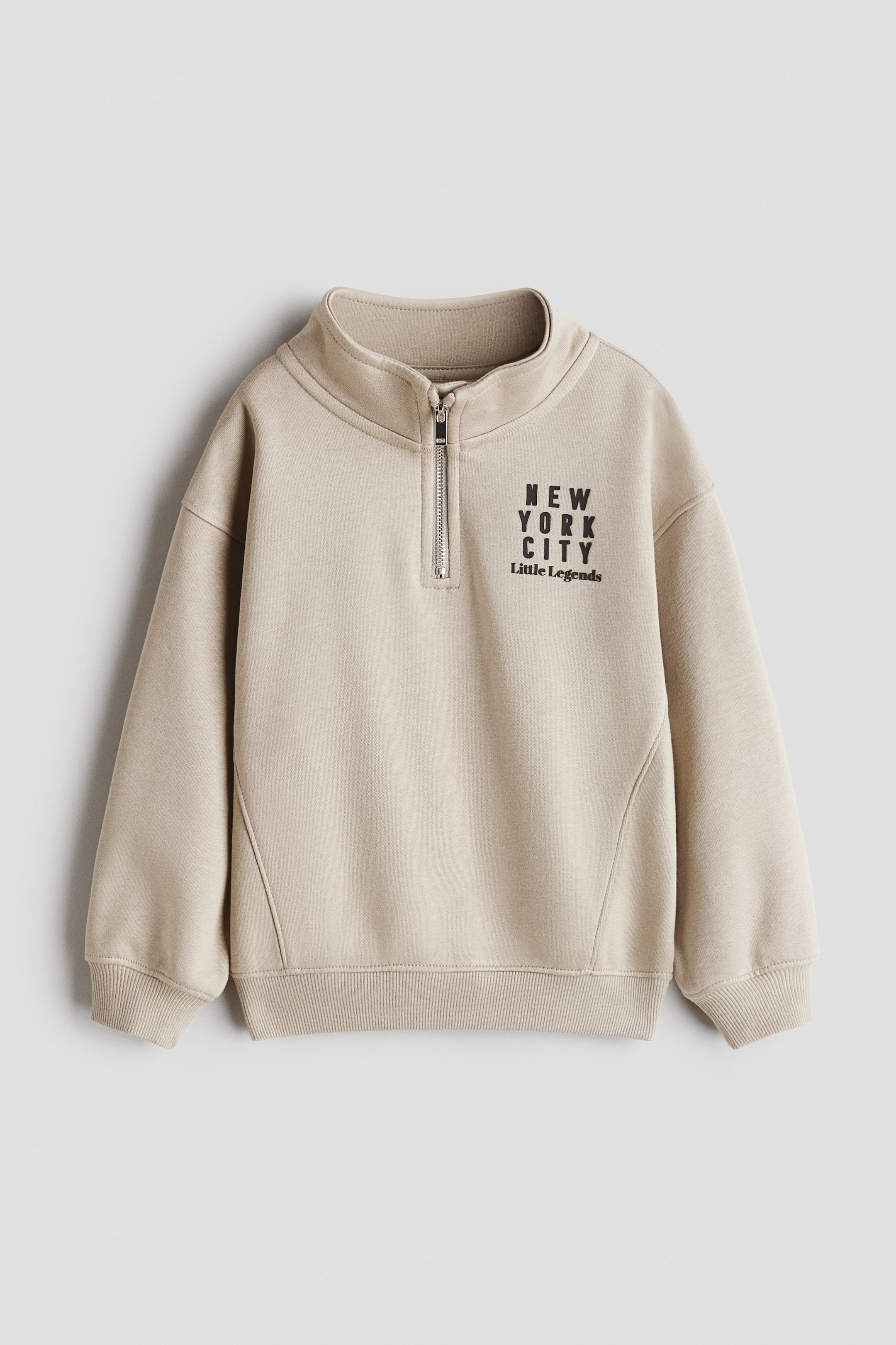 Büyük resmi görüntüle: Bej renkli, bol kesimli polar sweatshirt önden görünmektedir; üst kısmında fermuarlı, jarse astarlı dik yaka, ön kısmında "NEW YORK CITY Little Legends" motifi, düşük omuzlar ve ribanalı manşetler ile etek ucu bulunmaktadır.