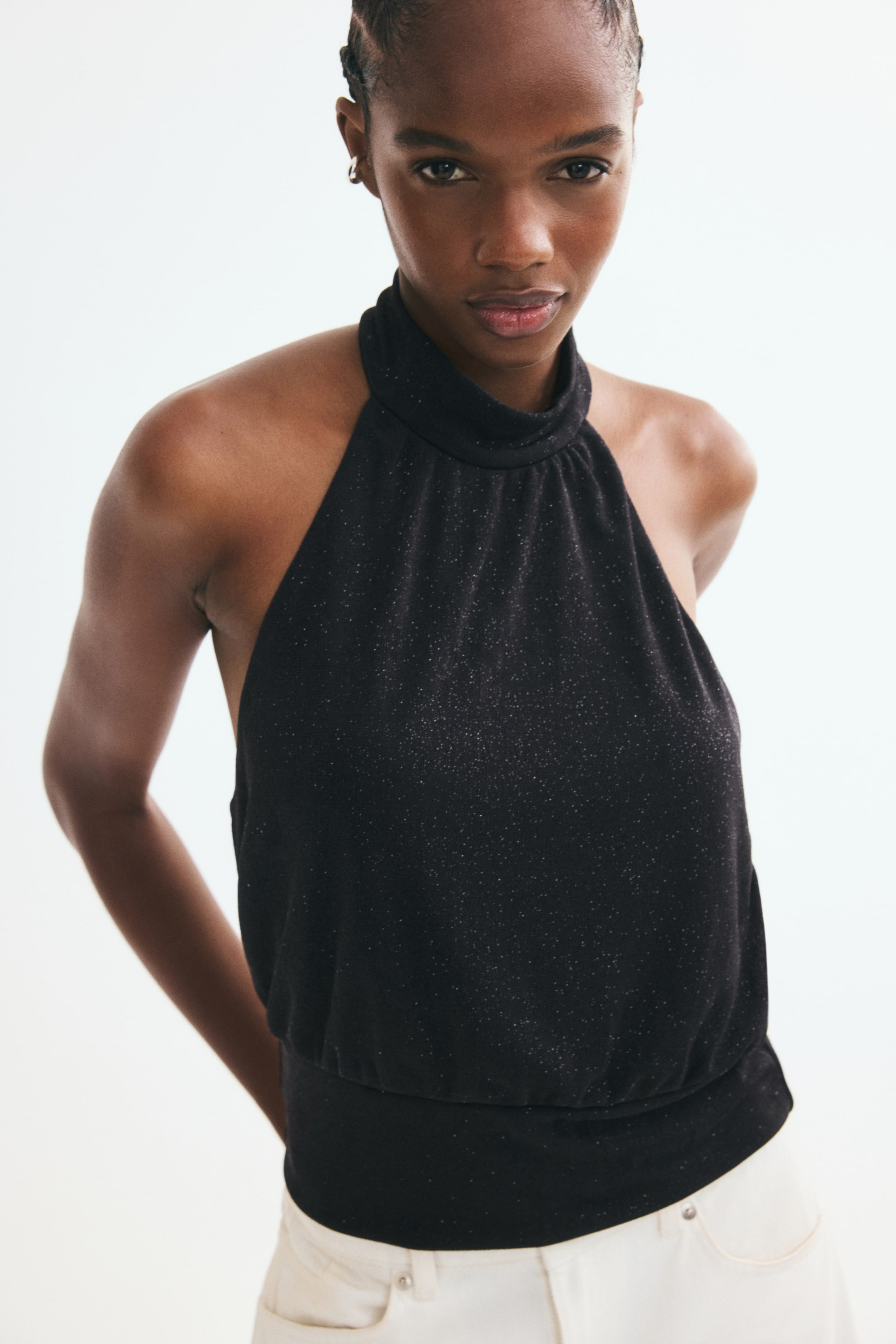 Größeres Bild ansehen: Ein schwarzes Neckholder-Top mit schimmernden Fäden wird von dem Model getragen und ist mit einem hohen, gerafften Ausschnitt sowie einem breiten, figurbetonten Bund gestaltet. Das Top ist mit hellen Hosen kombiniert.