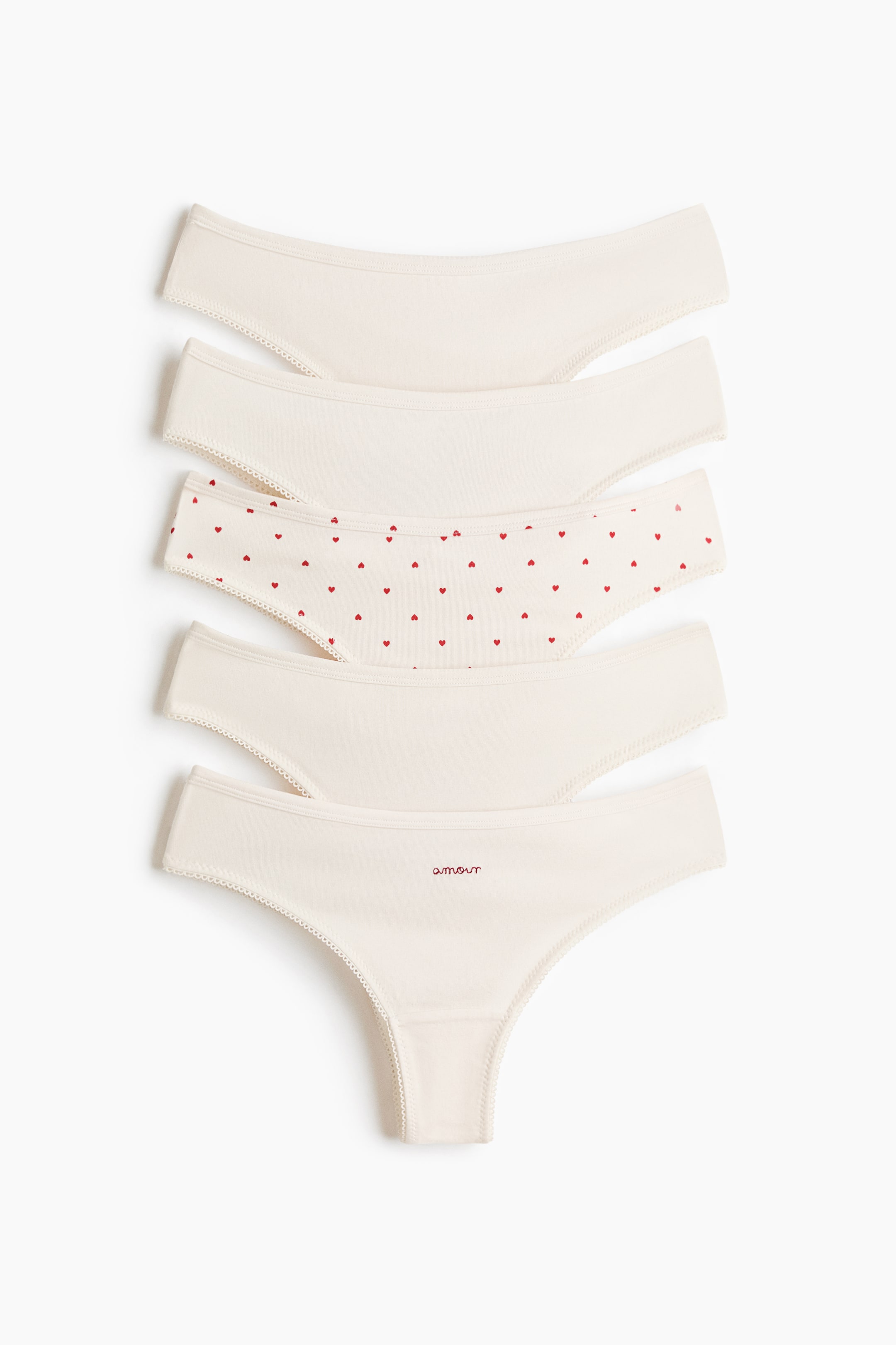 View larger image: Five pairs of cream-coloured underwear are lying stacked vertically, facing forward, with picot trim detailing their edges. The third pair is patterned with small red hearts, and the bottom thong-style pair is embroidered with "amour" in red.