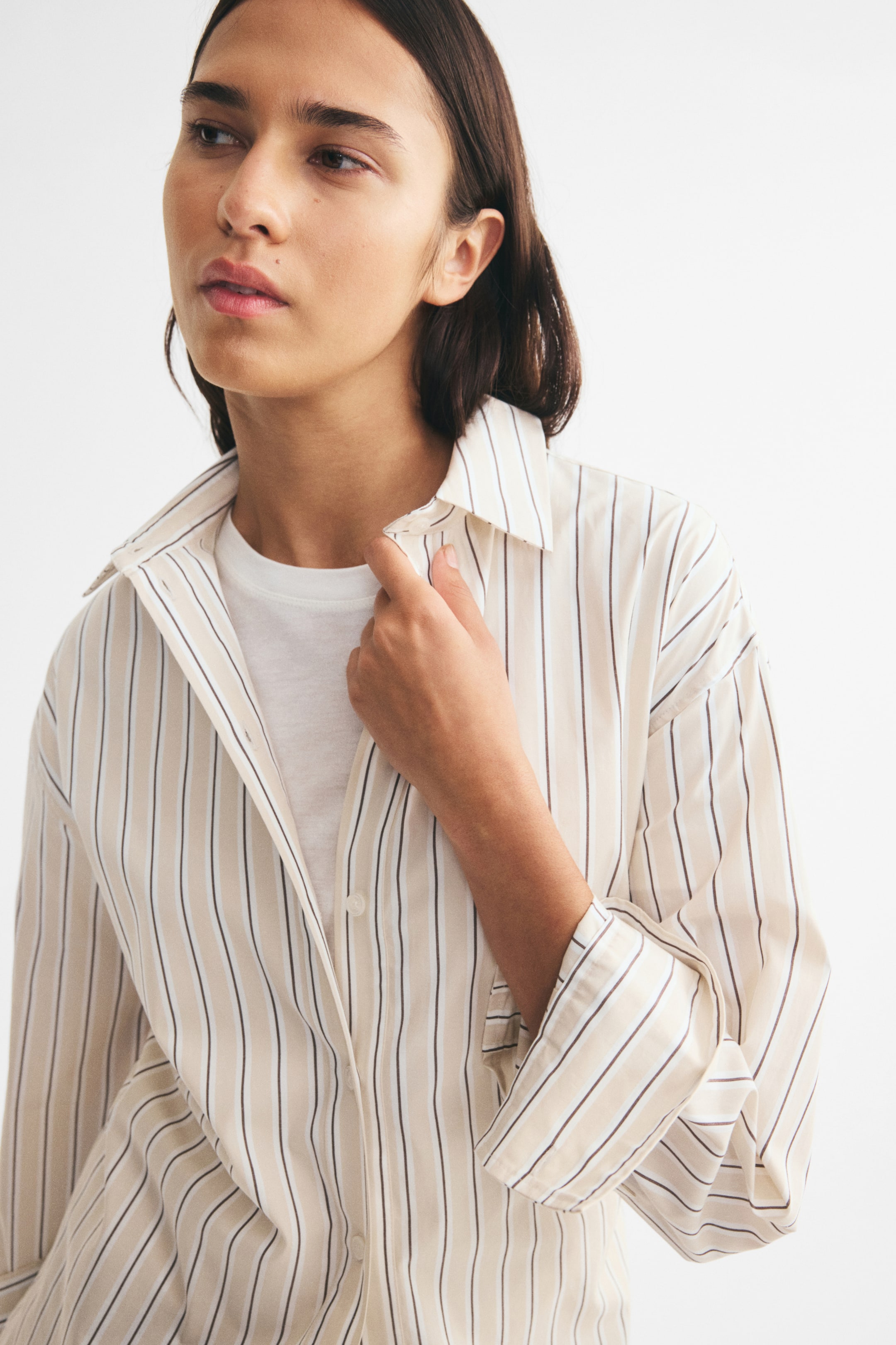 View larger image: An oversized cotton shirt with vertical beige, white, and dark stripes is being worn by the model over a white crew-neck t-shirt. The shirt features dropped shoulders and a collar, which the model is holding slightly open with one hand.