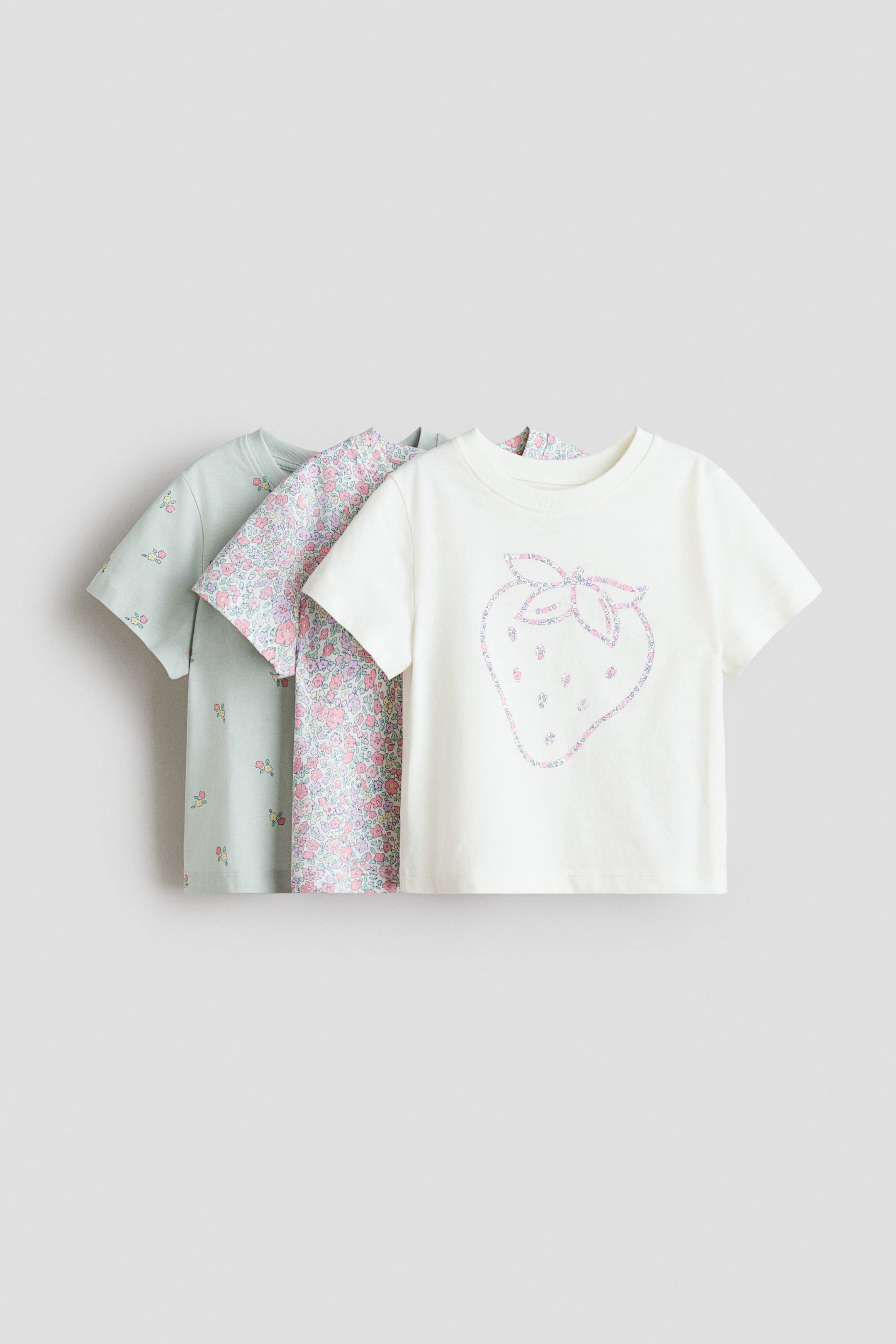 Büyük resmi görüntüle: Ön tarafta katmanlı olarak sergilenen yumuşak, baskılı pamuklu jarse kumaştan üçlü tişört seti. Her tişört, yaka çevresinde nervürlü bir şerit ve hafifçe düşürülmüş omuzlarla tasarlanmıştır, baskılar ise çeşitlilik göstermektedir: en öndeki tişört, büyük bir çilek kontürü baskısıyla kırık beyaz, ortadaki tişört ise küçük çiçek desenleriyle kaplıdır ve en arkadaki tişört, dağınık küçük çiçek desenleriyle açık yeşildir.