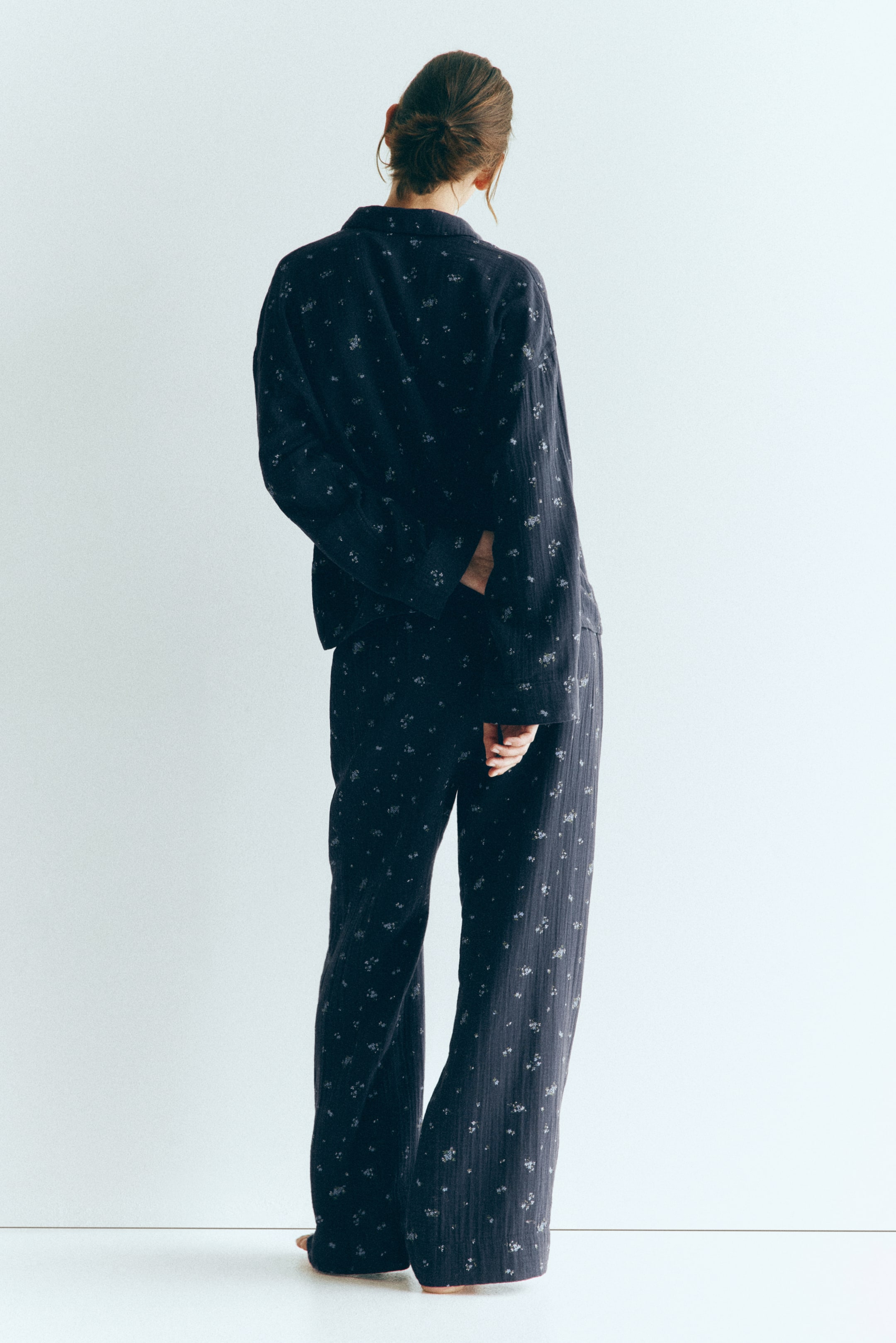View larger image: A dark-coloured, floral-patterned cotton muslin pyjama set, consisting of a long-sleeved shirt with dropped shoulders and matching straight-leg trousers, is being worn by the model. The model is standing with their back to the camera, showcasing the relaxed fit and soft, crinkled texture of the garments.