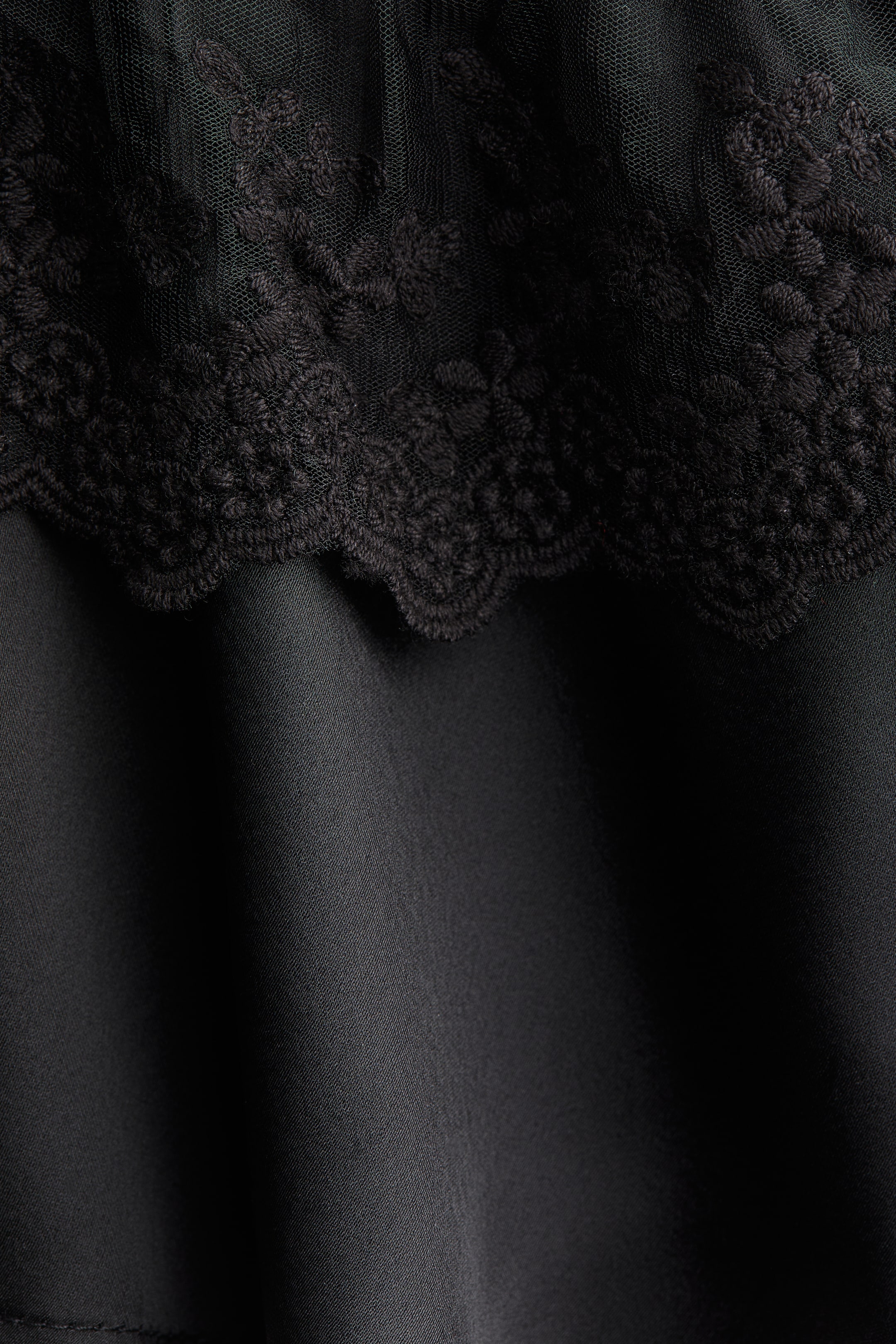 Ampliar la imagen: Una prenda negra presenta una sección superior de encaje transparente con intrincados bordados florales. El encaje muestra un borde festoneado que se encuentra con una tela negra lisa y sólida debajo.