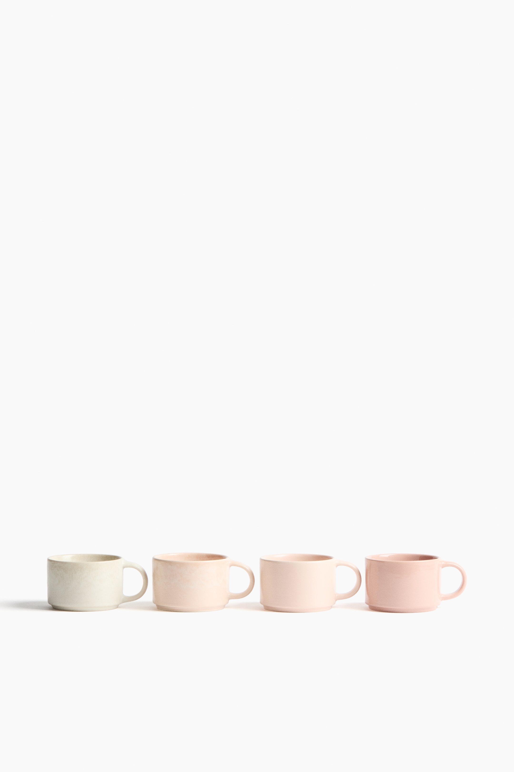 View larger image: Four stackable stoneware mugs are displayed in a row, each mug featuring a distinct glaze transitioning from a light greige to various shades of soft pink. Each mug features a C-shaped handle and a subtle ridge near its base indicating its stackable design.