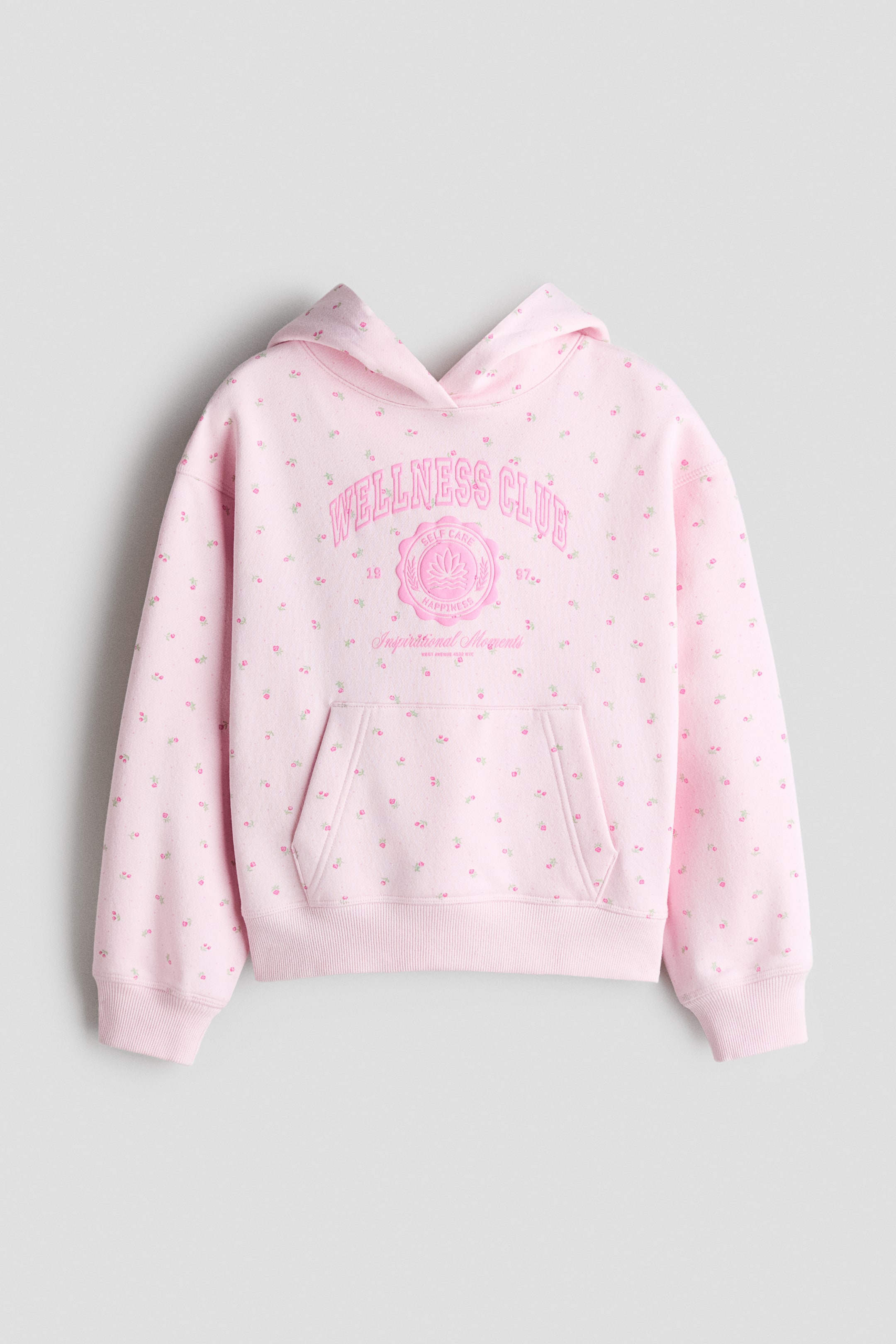 Se større billede: En lyserød oversized hoodie set forfra viser et heldækkende mini-blomstermønster og en central grafik med teksten "WELLNESS CLUB" over et cirkulært emblem. Hoodien har en wrap-front hætte, sænkede skuldre, en kængurulomme samt brede ribkanter ved manchetterne og kanten.