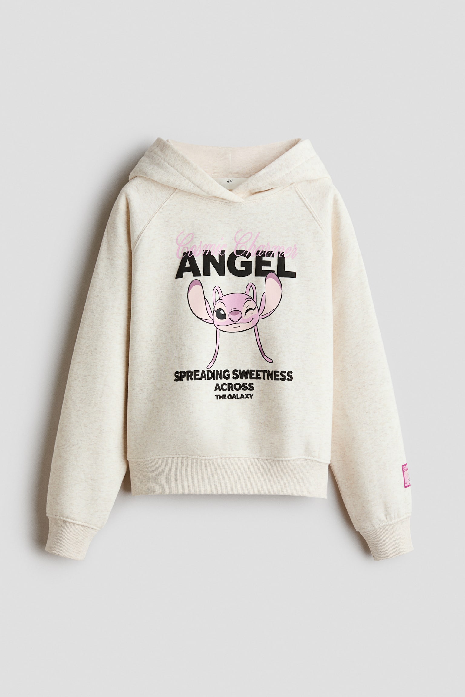 An oversized hoodie in a light beige brushed fleece fabric is shown front-facing, featuring a hood with a wrap-front design and long raglan sleeves. The chest area features a printed graphic of a pink cartoon character with text, while wide ribbed trims finish the cuffs and hem. - 1