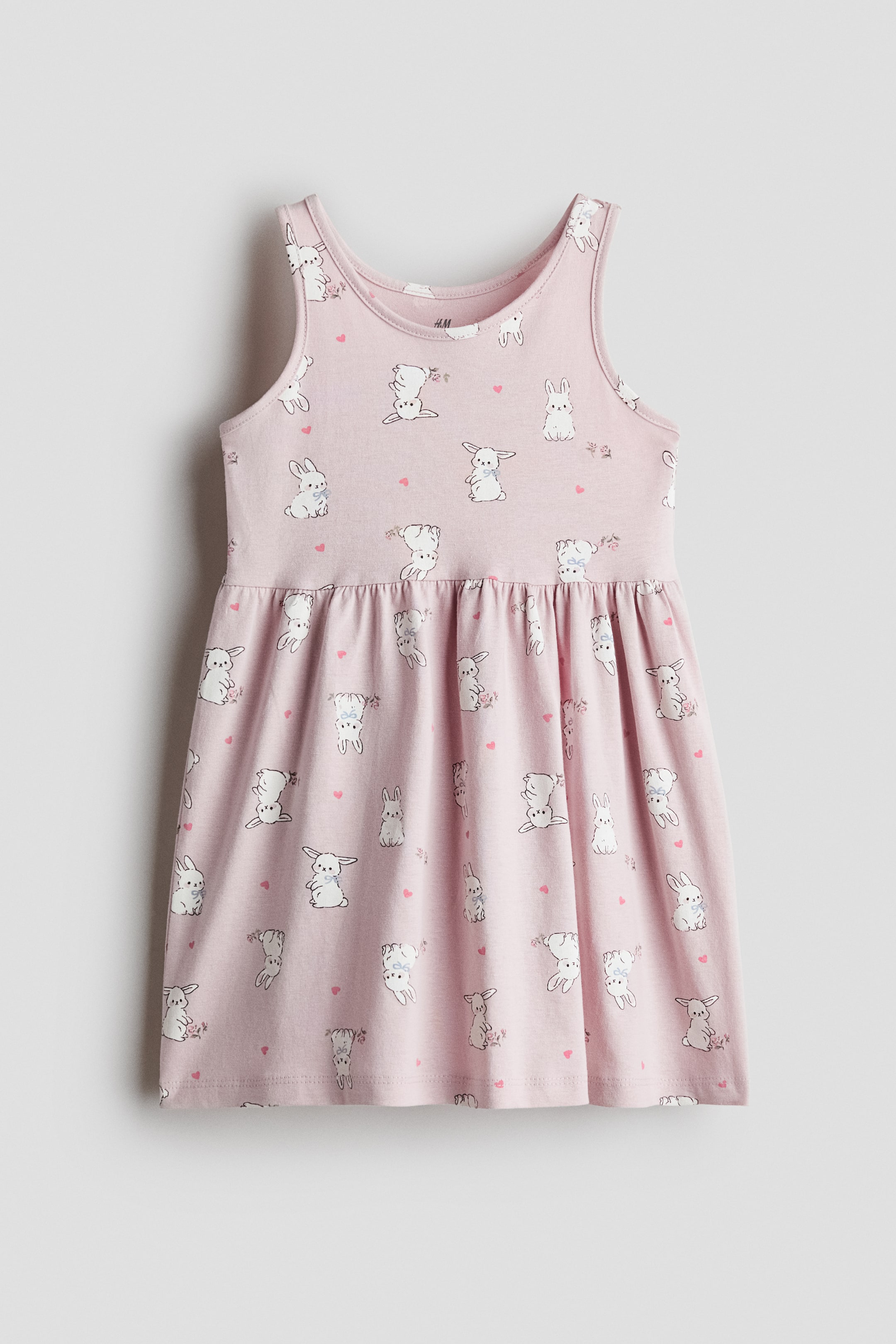View larger image: A light pink sleeveless jersey dress is shown in a front-facing view, featuring an all-over print of small white rabbits and tiny pink hearts. The dress has a round neckline, a gathered seam at the waist, and a flared skirt.