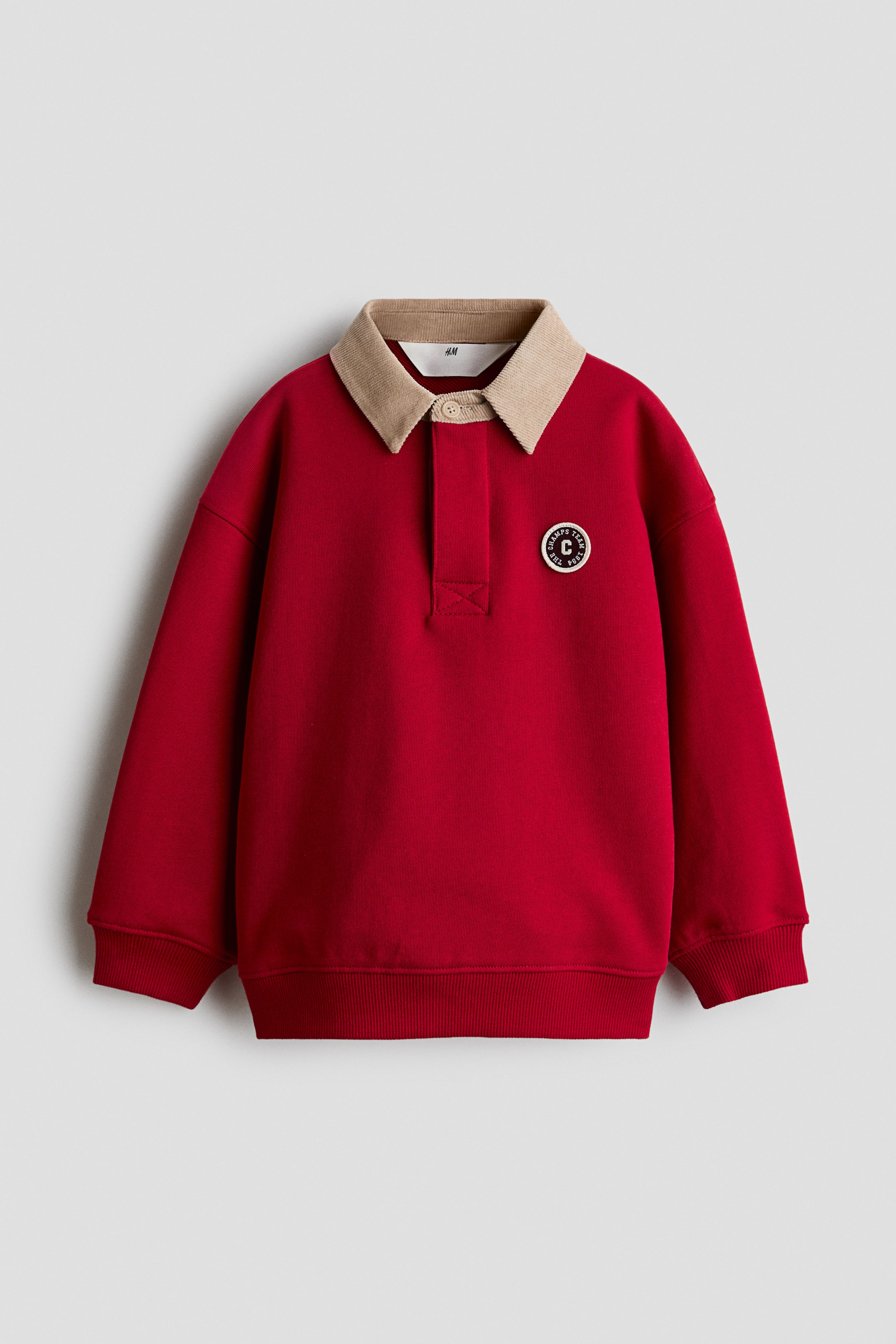 View larger image: A red rugby shirt is shown from the front, featuring a beige corduroy collar, a button placket, and a circular embroidered patch on the left chest. The shirt has dropped shoulders, long sleeves with ribbed cuffs, and a ribbed hem.
