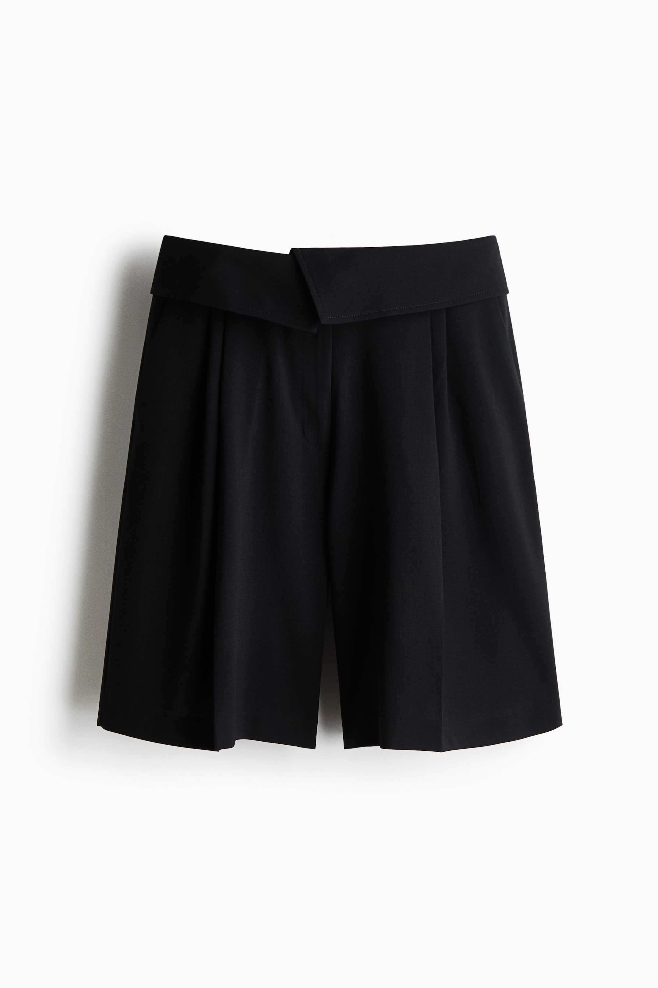 View larger image: Black shorts showcase a front design with a wide, overlapping waistband that forms a V-cut at the front. They feature prominent front pleats and wide legs, extending to knee-length.