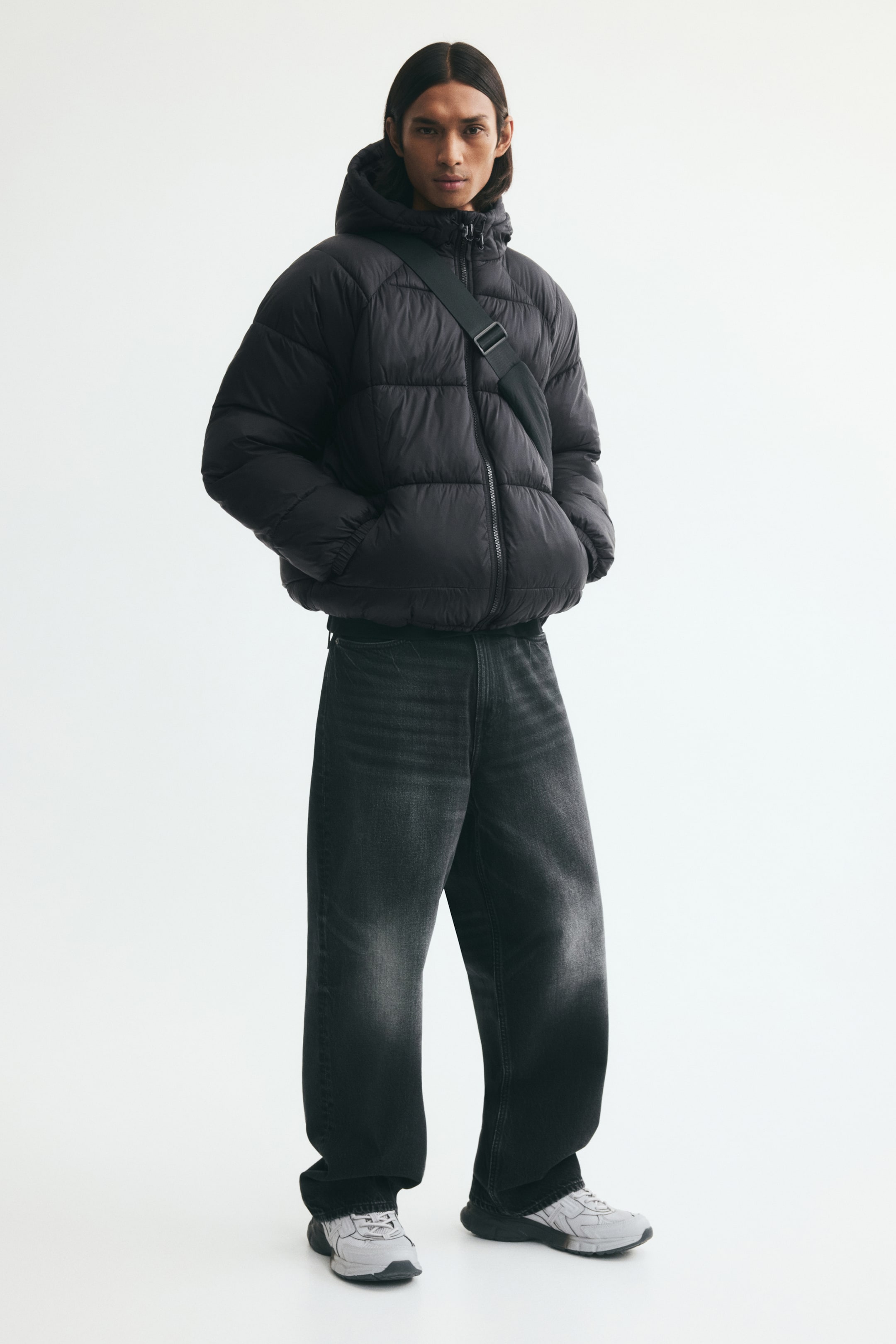 View larger image: A black padded puffer jacket featuring a quilted weave, a stand collar, and a hood is being worn by the model. The model is standing with hands in the front pockets, styling the jacket with wide-leg grey jeans and a black crossbody strap.
