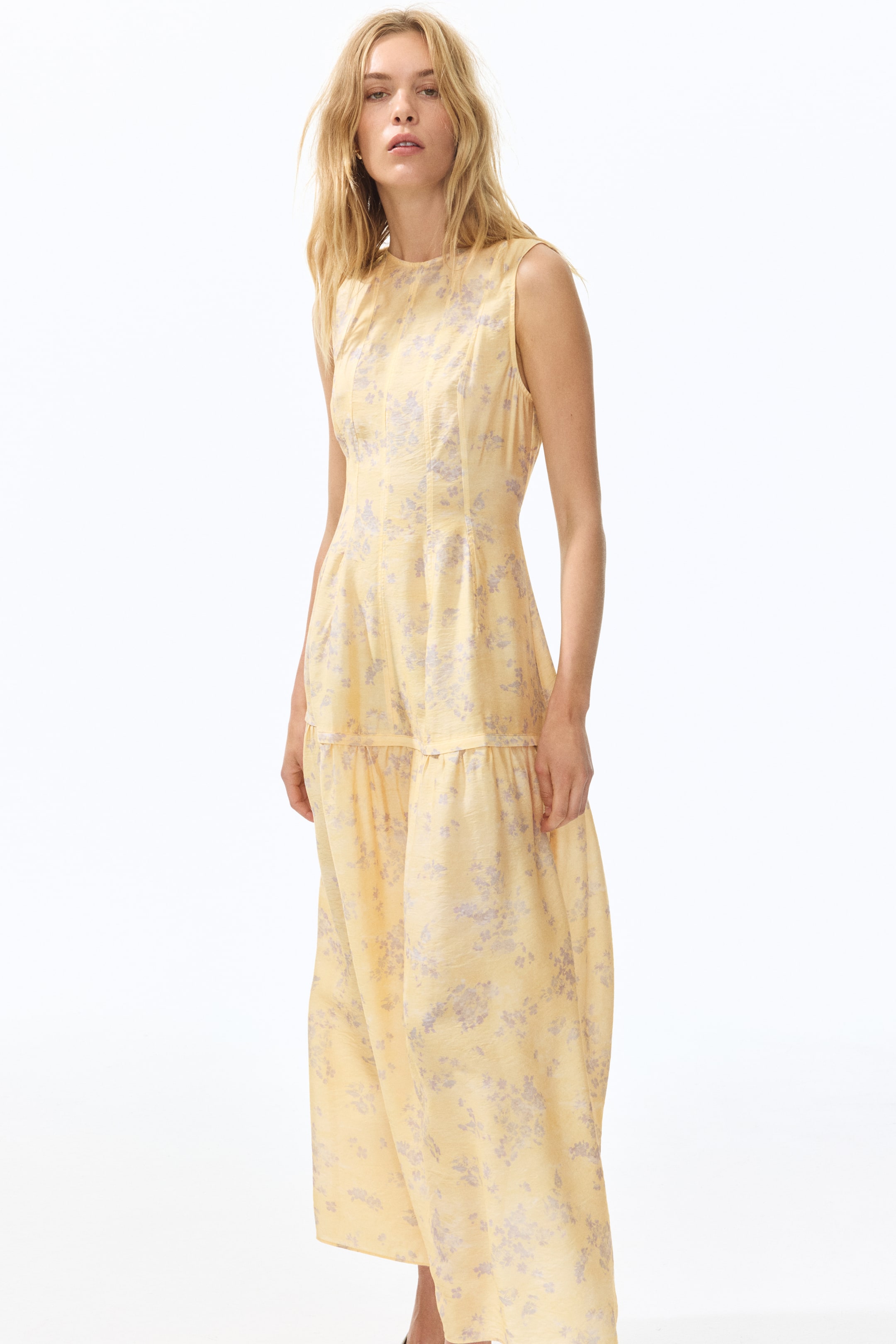 View larger image: Presenting a vibrant yellow sleeveless maxi dress adorned with a delicate light purple floral pattern. This elegant piece showcases a round neckline and a fitted bodice accentuated by vertical seaming. The tiered skirt, with its gentle gathering, is subtly angled to the right, adding a touch of sophistication to the overall design.