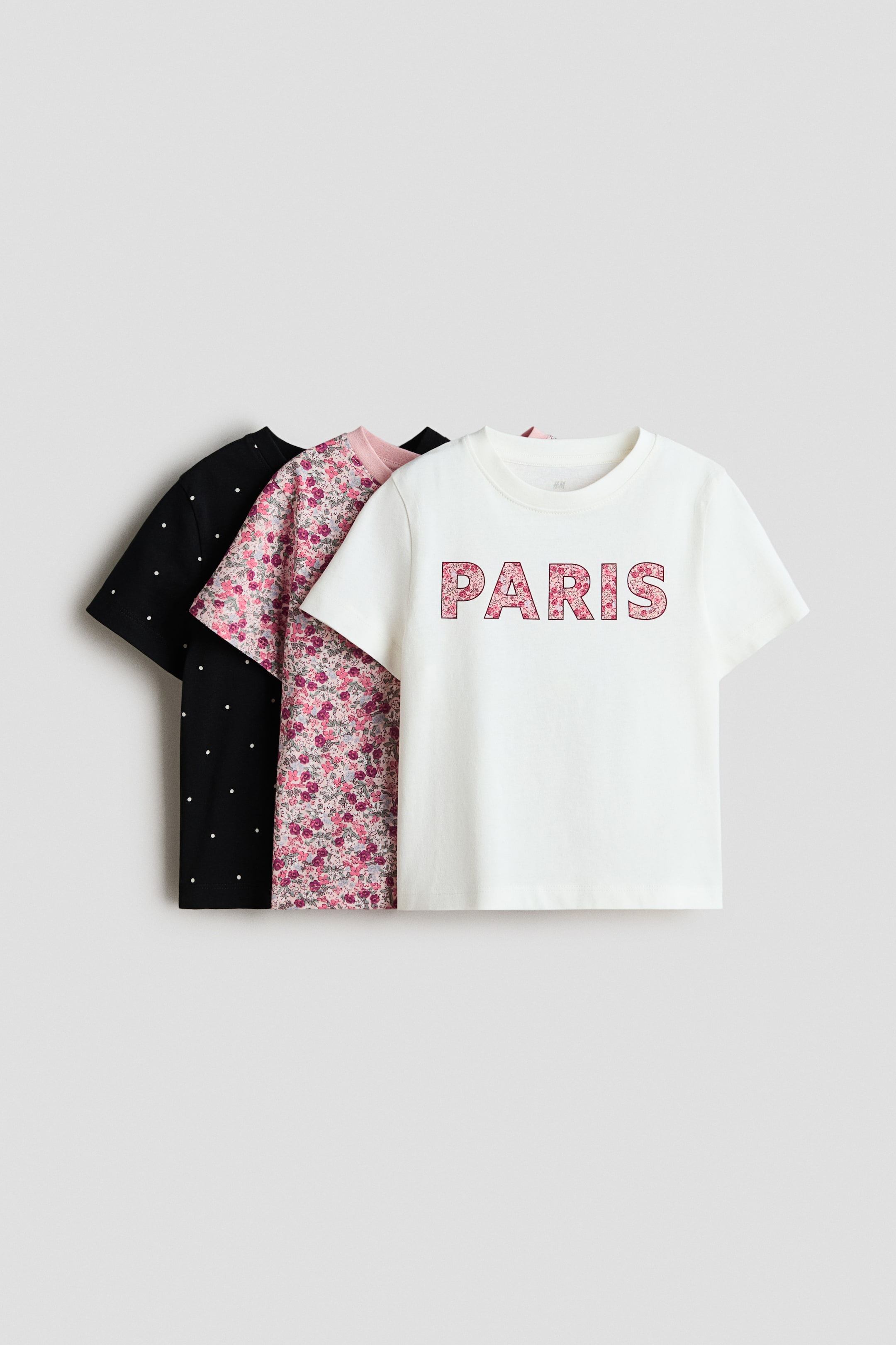 Voir une image plus grande: Un ensemble de trois t-shirts à manches courtes et à col rond est présenté, incliné de face, avec le t-shirt blanc à l'avant arborant "PARIS" dans un motif floral rose. Le t-shirt rose du milieu affiche un petit imprimé floral, tandis que le t-shirt noir à l'arrière présente des pois blancs.