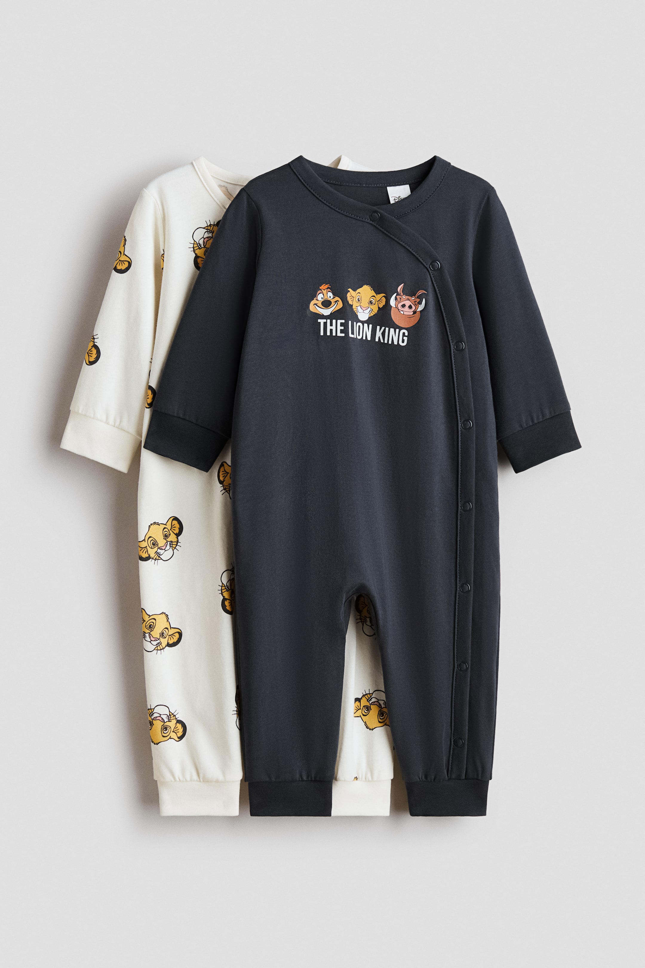 Grotere afbeelding bekijken: Twee all-in-one pyjama's met lange mouwen van bedrukte katoenen jersey worden gepresenteerd, één donkergrijs en één crèmekleurig. De donkergrijze pyjama op de voorgrond heeft een grafische print van 'THE LION KING'-figuren op de borst, diagonaal geplaatste drukknopen over de voorkant en langs één been, en zwarte geribbelde boorden en zomen, terwijl de crèmekleurige pyjama erachter een all-overprint toont van het gezicht van een leeuwtje en crèmekleurige geribbelde boorden en zomen heeft.
