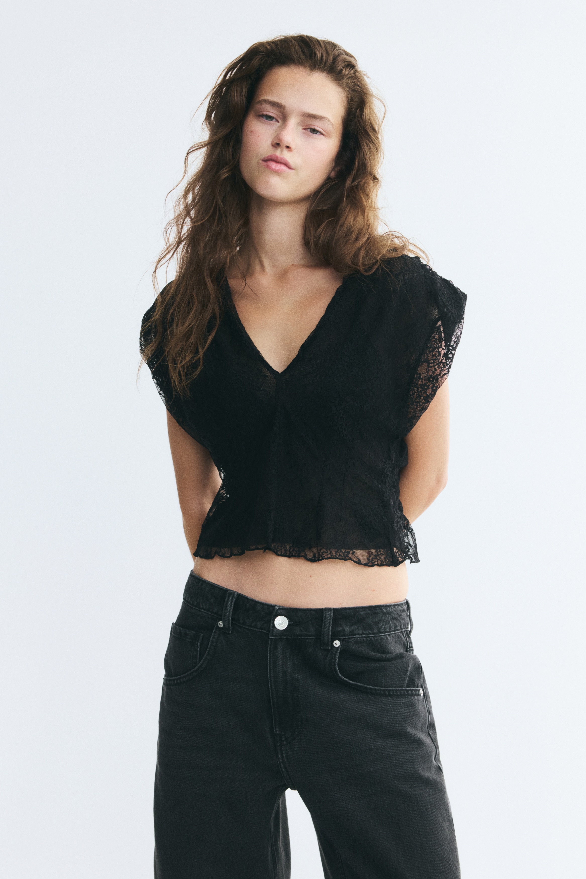 Un haut noir court sans manches en dentelle extensible est prÃ©sentÃ© de face, avec un profond dÃ©colletÃ© en V, des Ã©paules rembourrÃ©es avec dÃ©tails froncÃ©s et des pinces de mise en forme le long du corsage. Le haut prÃ©sente un ourlet droit et une construction partiellement doublÃ©e, laissant les Ã©paules et le haut de la poitrine en transparence.