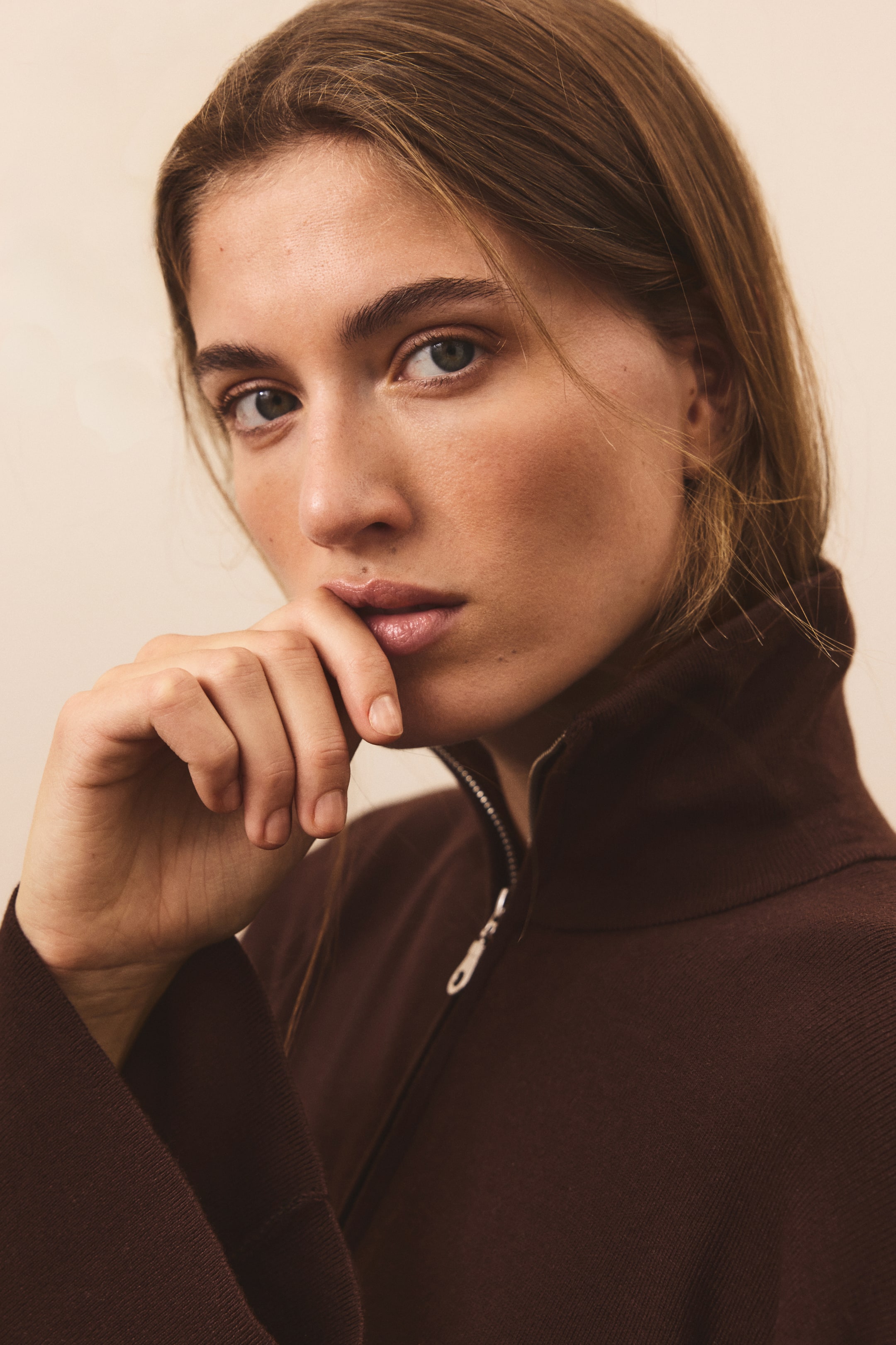 Grösseres Bild ansehen: Eine Nahaufnahme von vorne zeigt einen dunkelbraunen Feinstrickpullover mit hohem Stehkragen und silbernem Half-Zip-Verschluss. Der Pullover wird leicht geöffnet getragen und hebt so die weiche Struktur sowie den markanten Ausschnitt hervor.