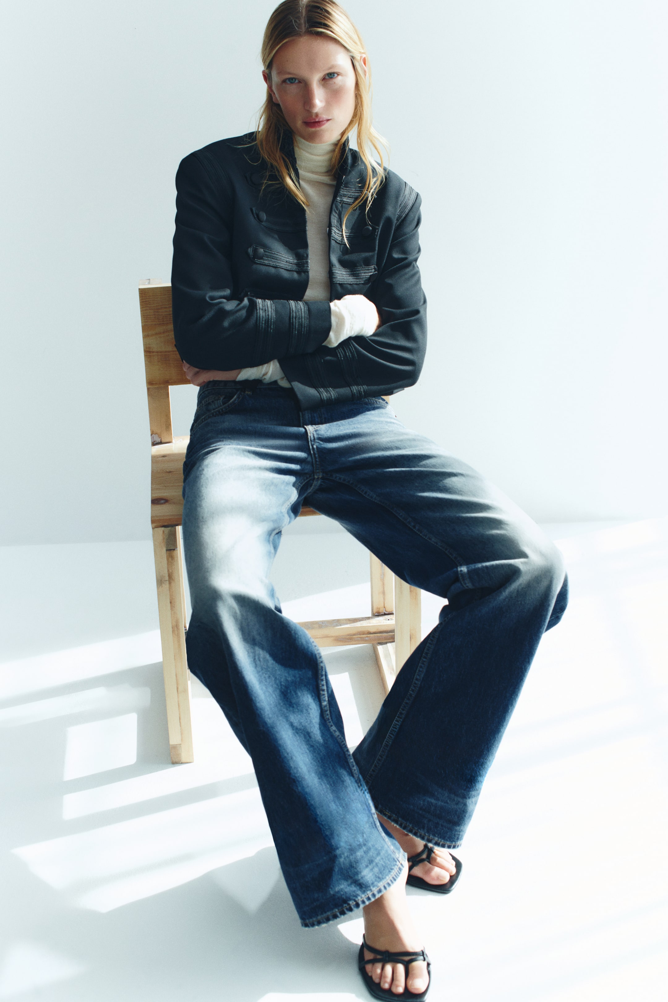 View larger image: Wide-leg blue denim jeans with a regular waist and faded thighs are being worn by the model, who is sitting on a wooden chair. The jeans are styled with a cream roll-neck, a black structured jacket, and black strappy sandals.